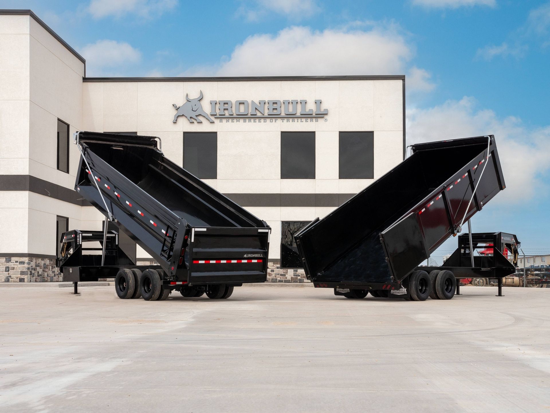 Two black Ironbull dump trailers in a paved lot, one with the bed raised.
