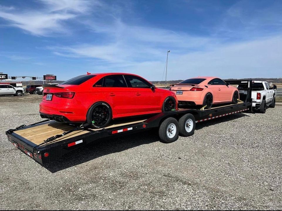 Two cars, red sedan and peach coupe, on a trailer being towed by a white truck on a gravel lot under a blue sky.