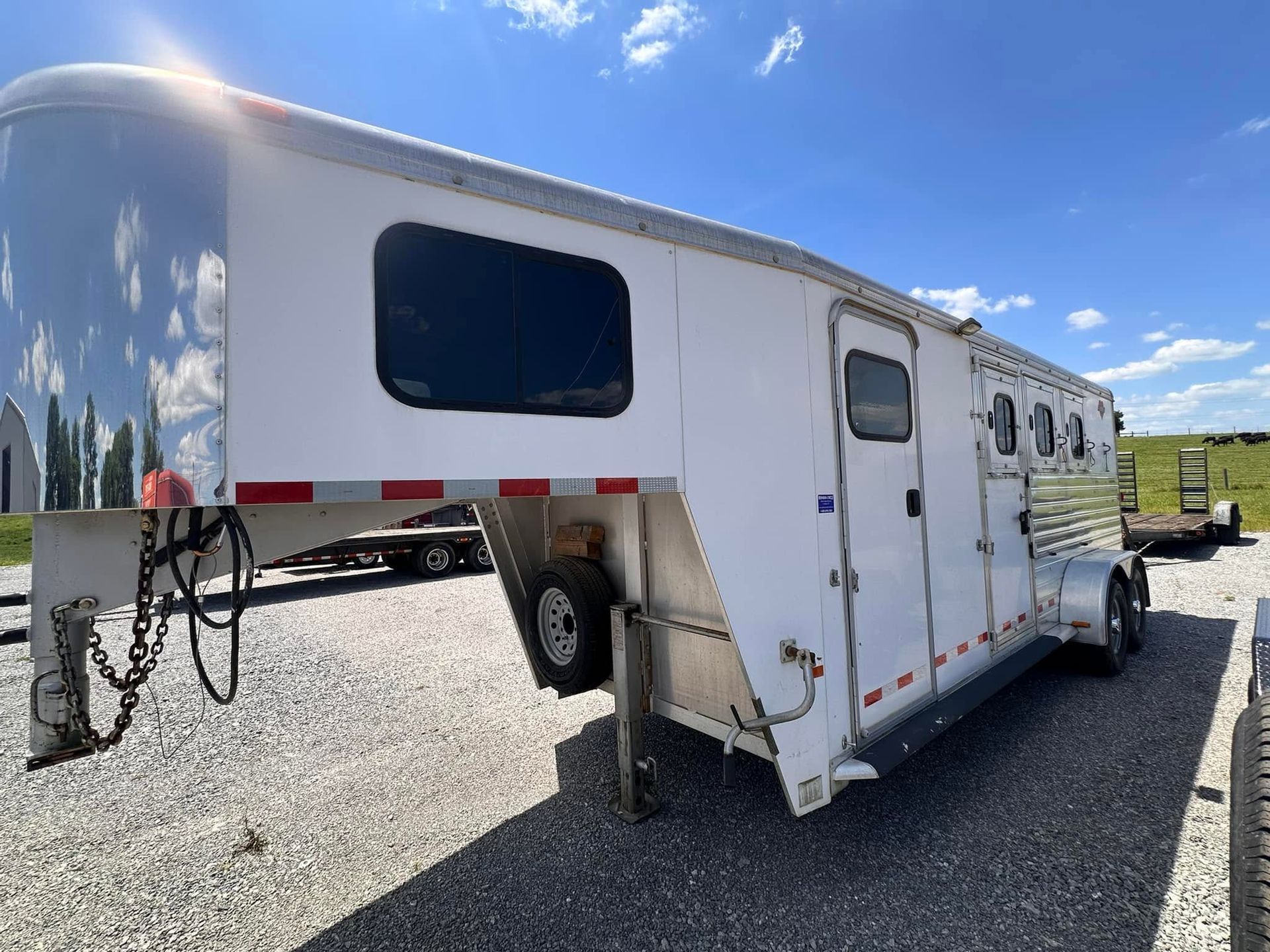 White horse trailer parked on gravel under a blue sky, hitched to a vehicle with a large black horse trailer beside it