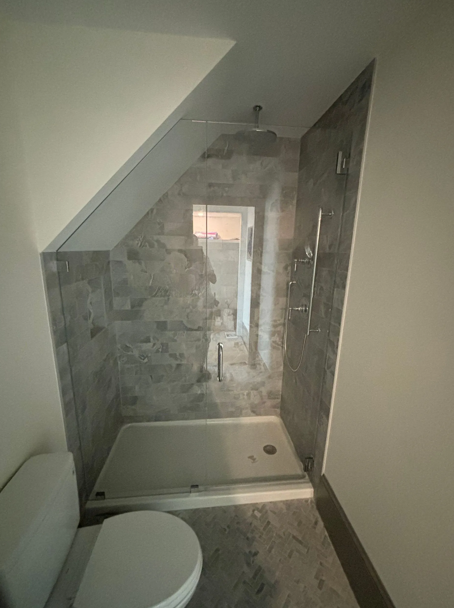 A bathroom with a toilet and a walk-in shower under a sloped ceiling. Gray tiled walls and floor.