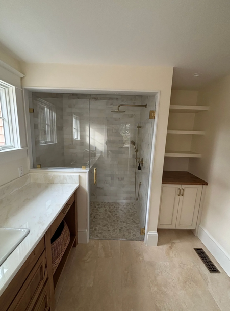 Bathroom with glass shower, marble counter, wooden vanity, and built-in shelves. Beige and gold tones.
