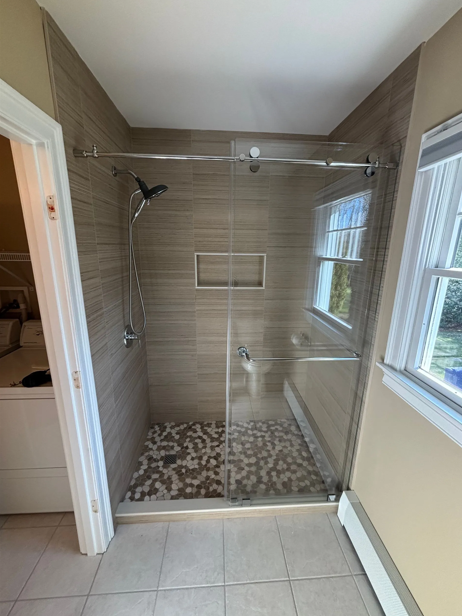 Shower with glass doors, tiled walls, and pebble floor. Two windows flank the shower.