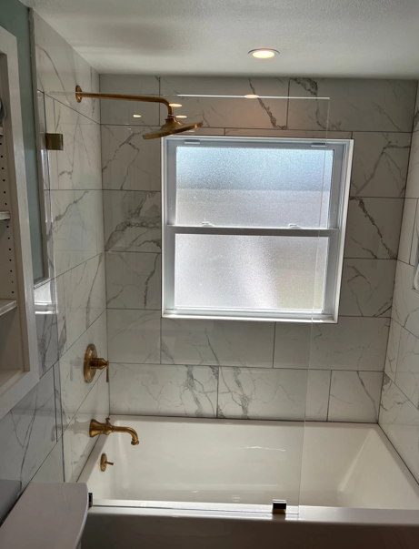Bathroom with a white tub, marble-like tiled walls, gold fixtures, and a window.