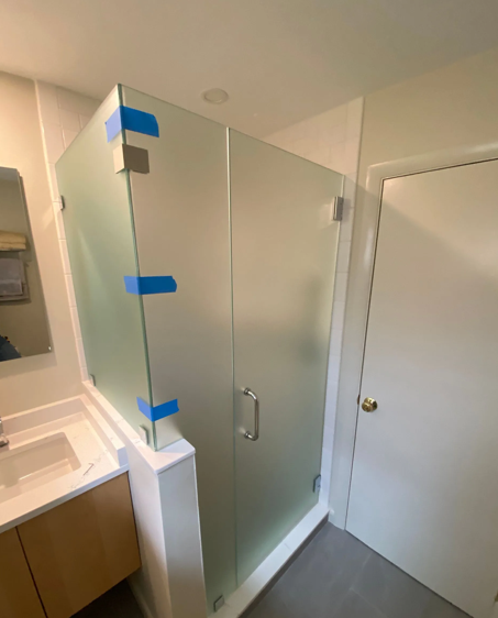 Frosted glass shower enclosure in a bathroom, featuring a door and corner panels, with blue tape on edges.