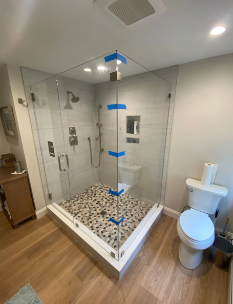 A newly renovated bathroom with a glass shower, pebble floor, and toilet on a hardwood floor.