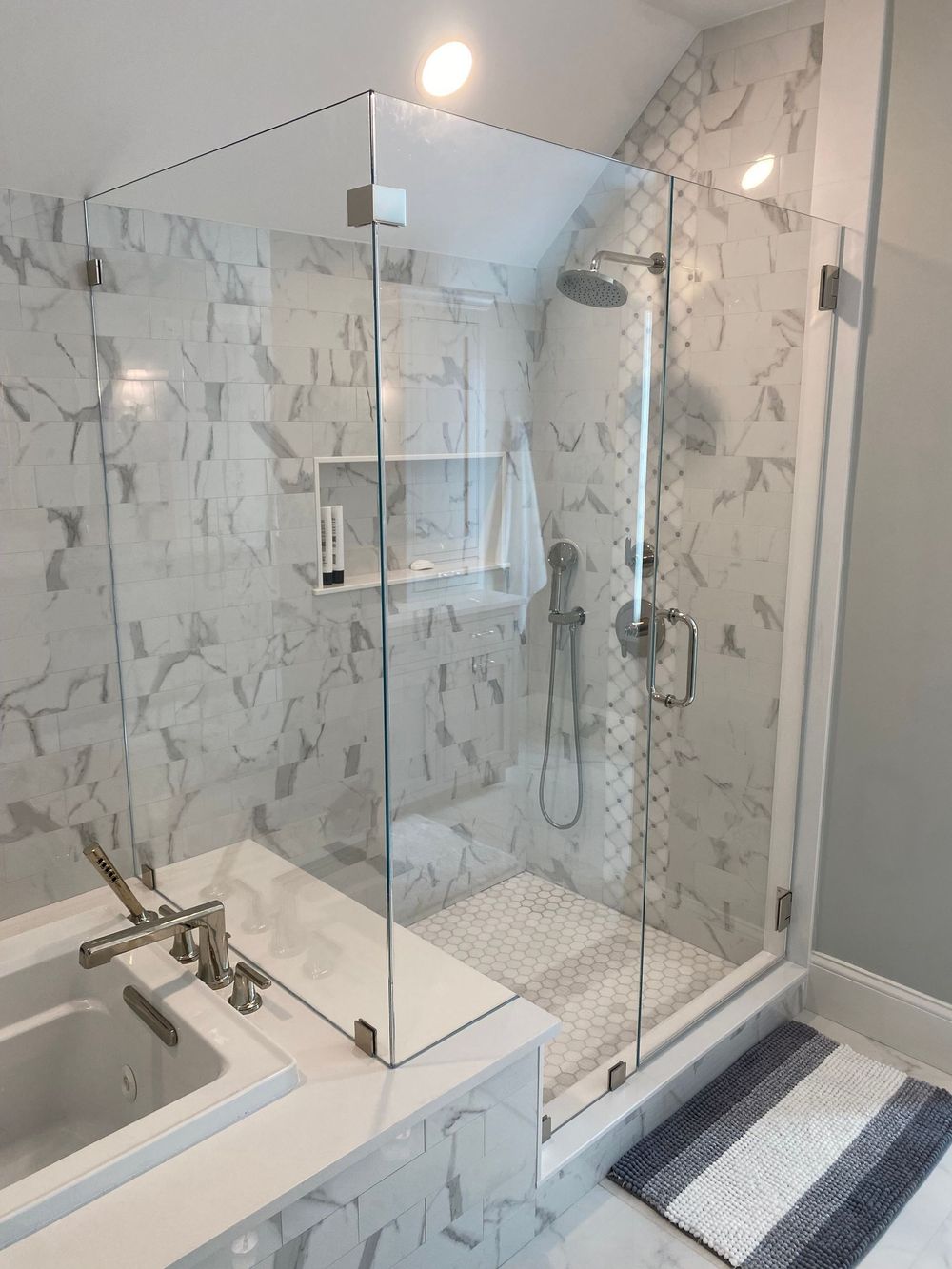 Glass-enclosed shower with white marble tile, a bench, and chrome fixtures. Gray and white bathmat.