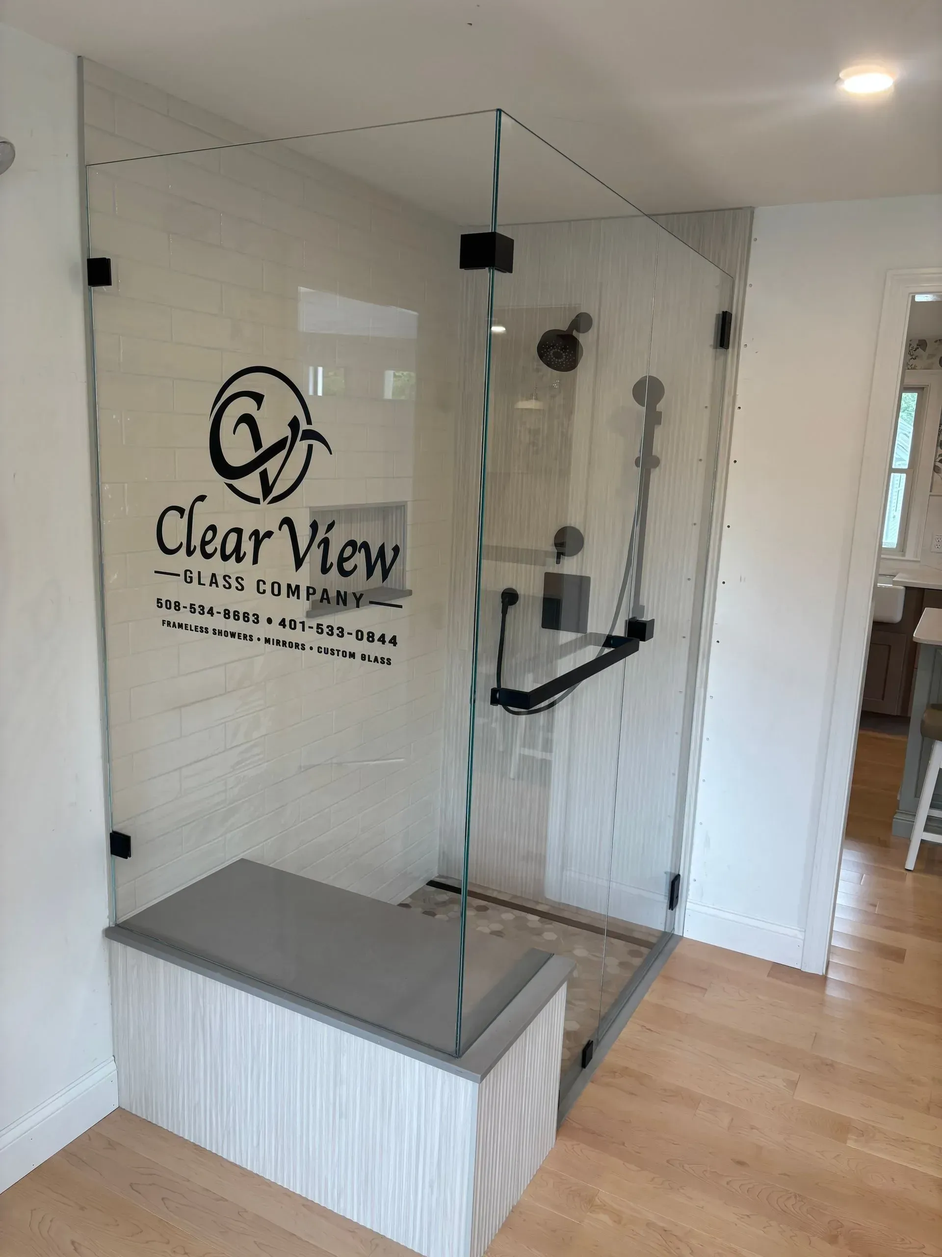 Glass shower enclosure with a built-in bench, black fixtures, and a logo on the glass.