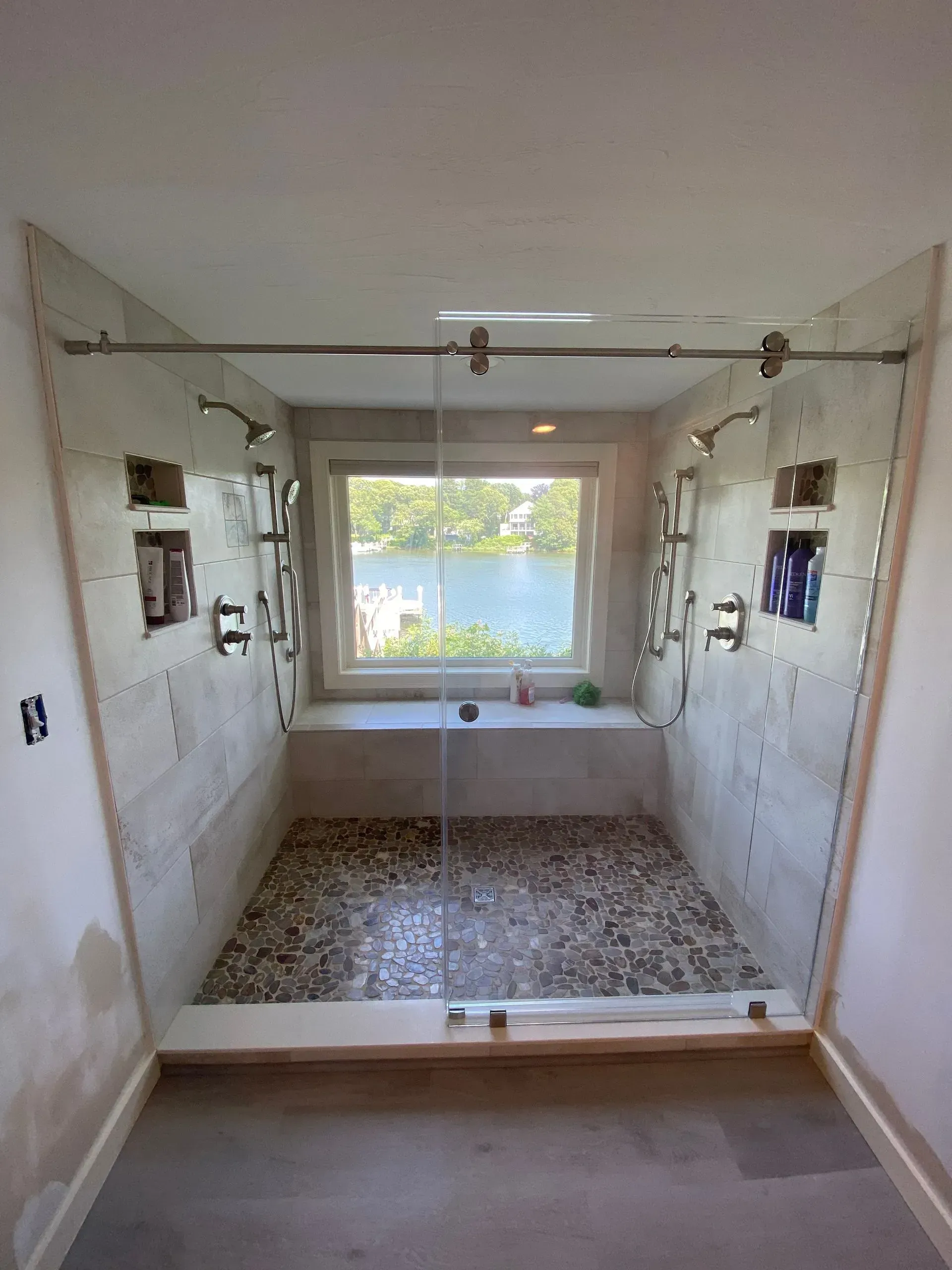 Spacious shower with pebble floor, glass doors, multiple showerheads, and a window overlooking a lake.
