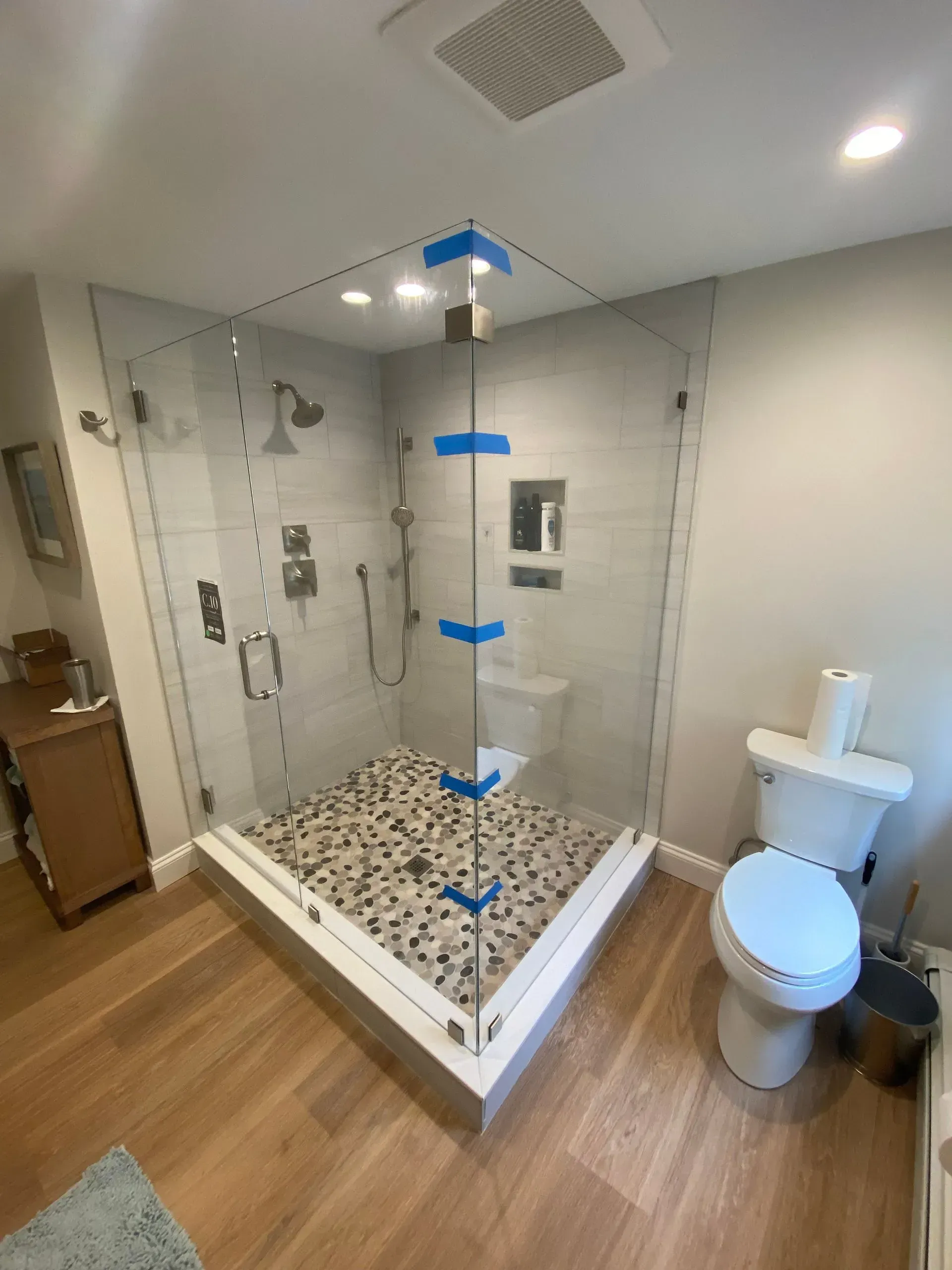 Corner shower with glass doors, pebble floor, toilet, and wooden floor. White and gray bathroom.