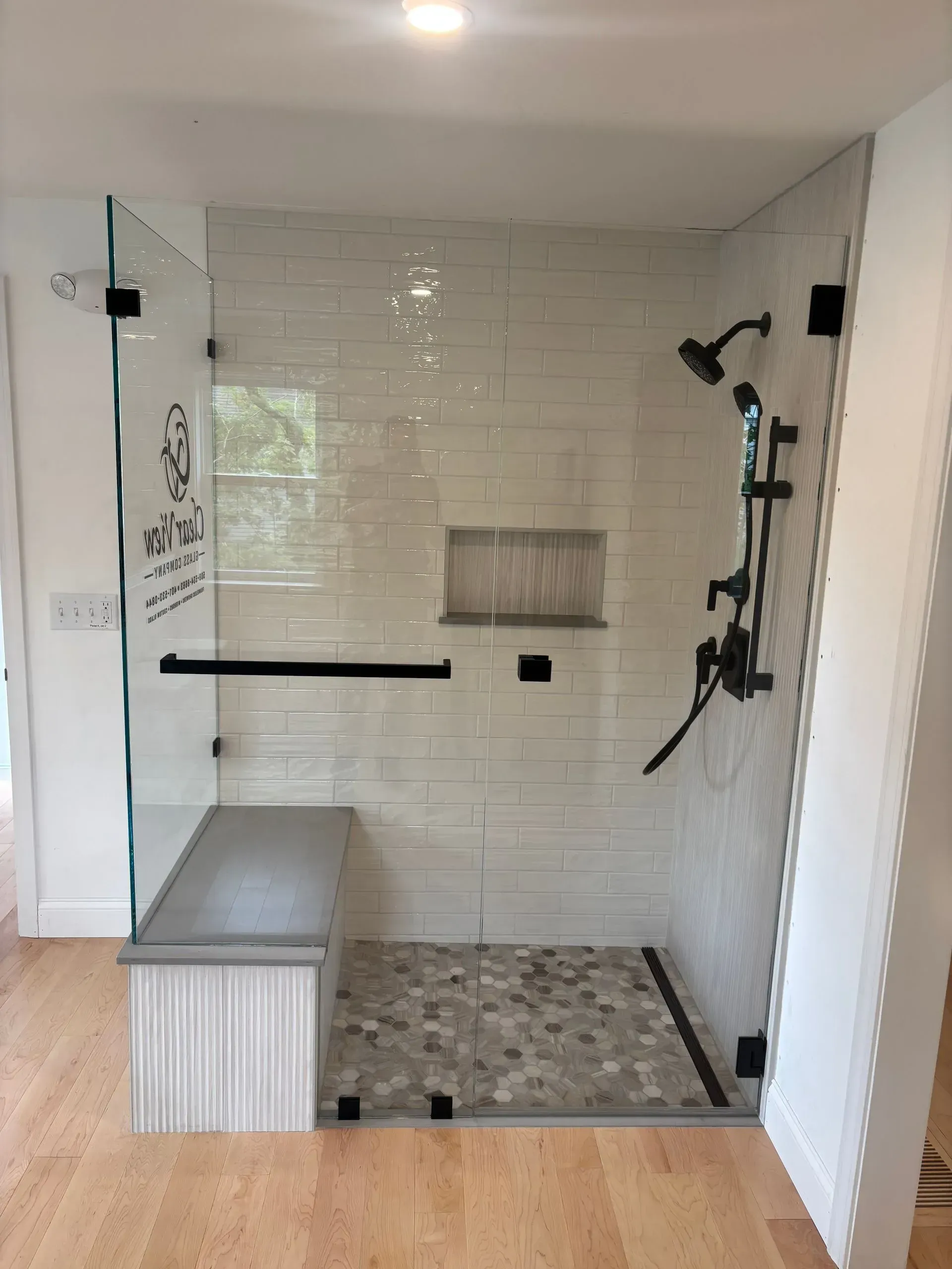 Accessible walk-in shower with bench and pebbled floor. Black fixtures, white tiled walls, and clear glass enclosure.