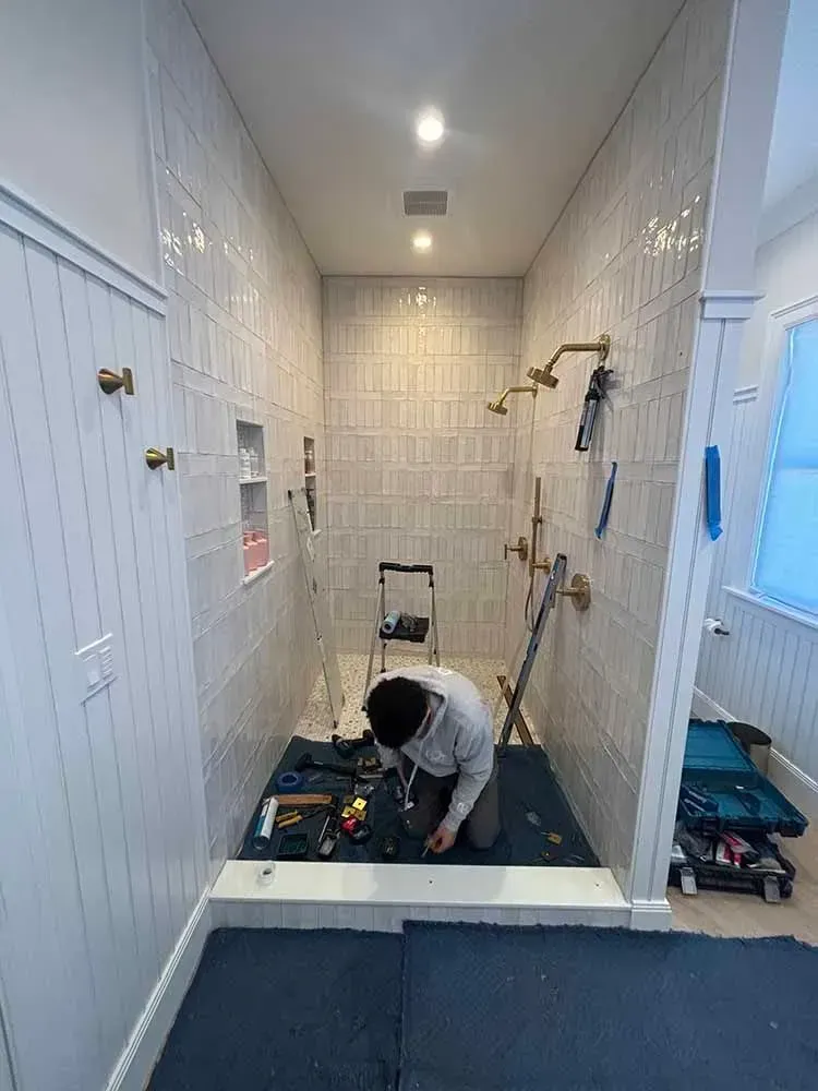 Person installing fixtures in a tiled shower, using tools on a blue mat. Brightly lit interior.