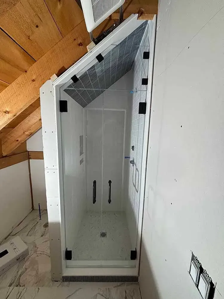 Shower stall built into sloped attic ceiling. Glass door, white walls, black hardware.