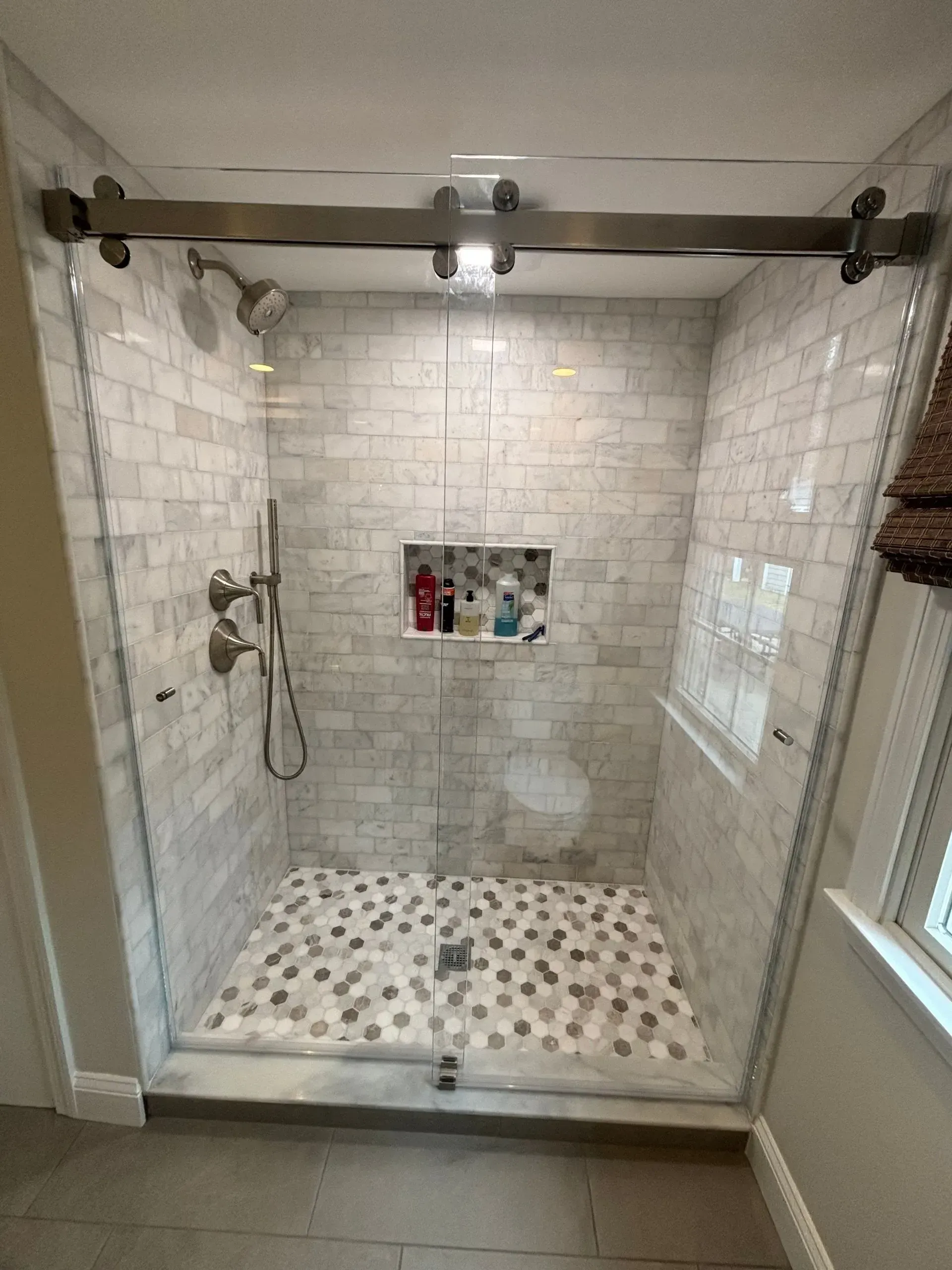 Glass-enclosed shower with a sliding door, gray tile, pebble floor, and built-in shelf.