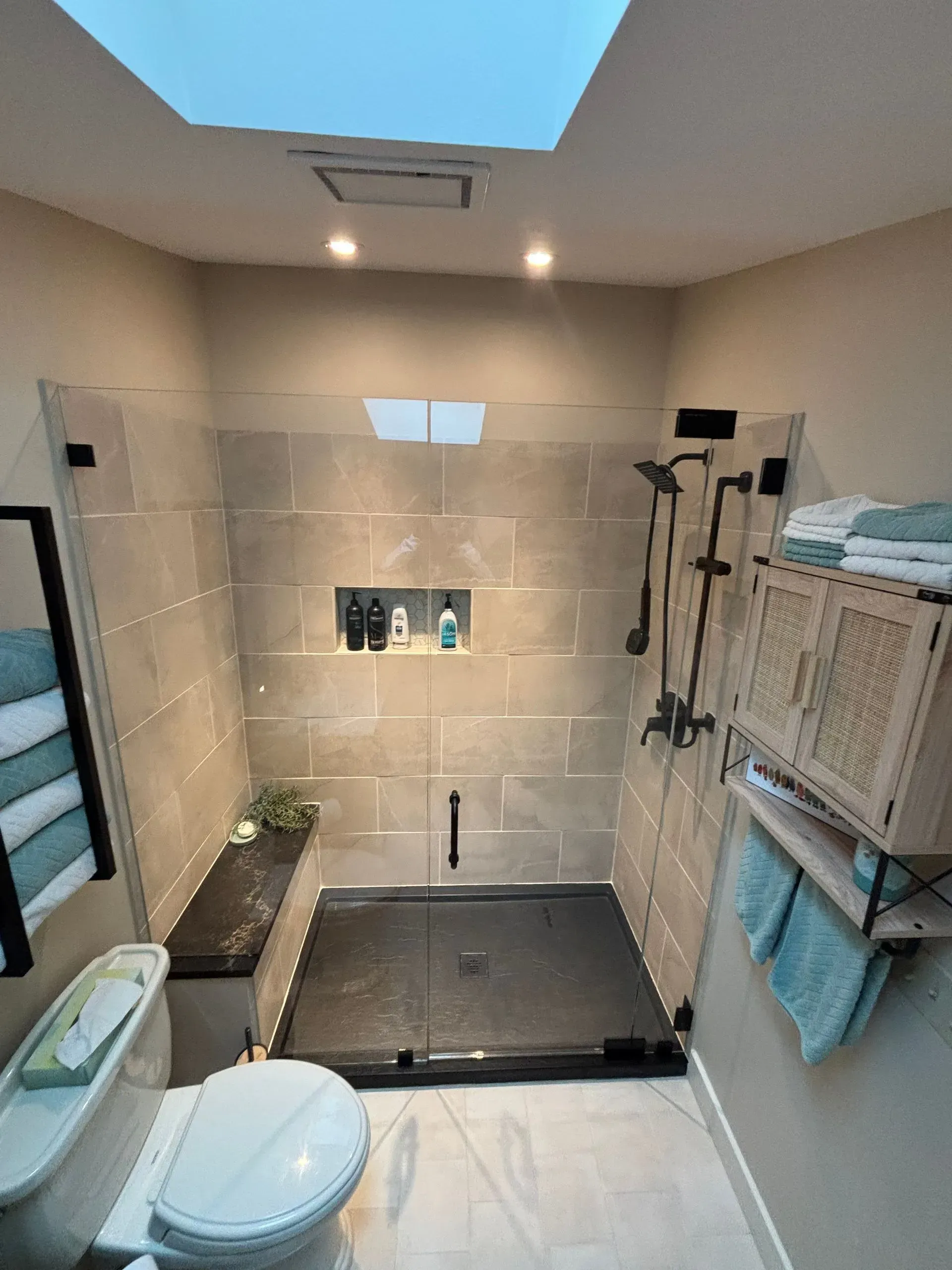 A modern bathroom with a large shower, toilet, and vanity. Light beige tiles and fixtures; light blue towels.