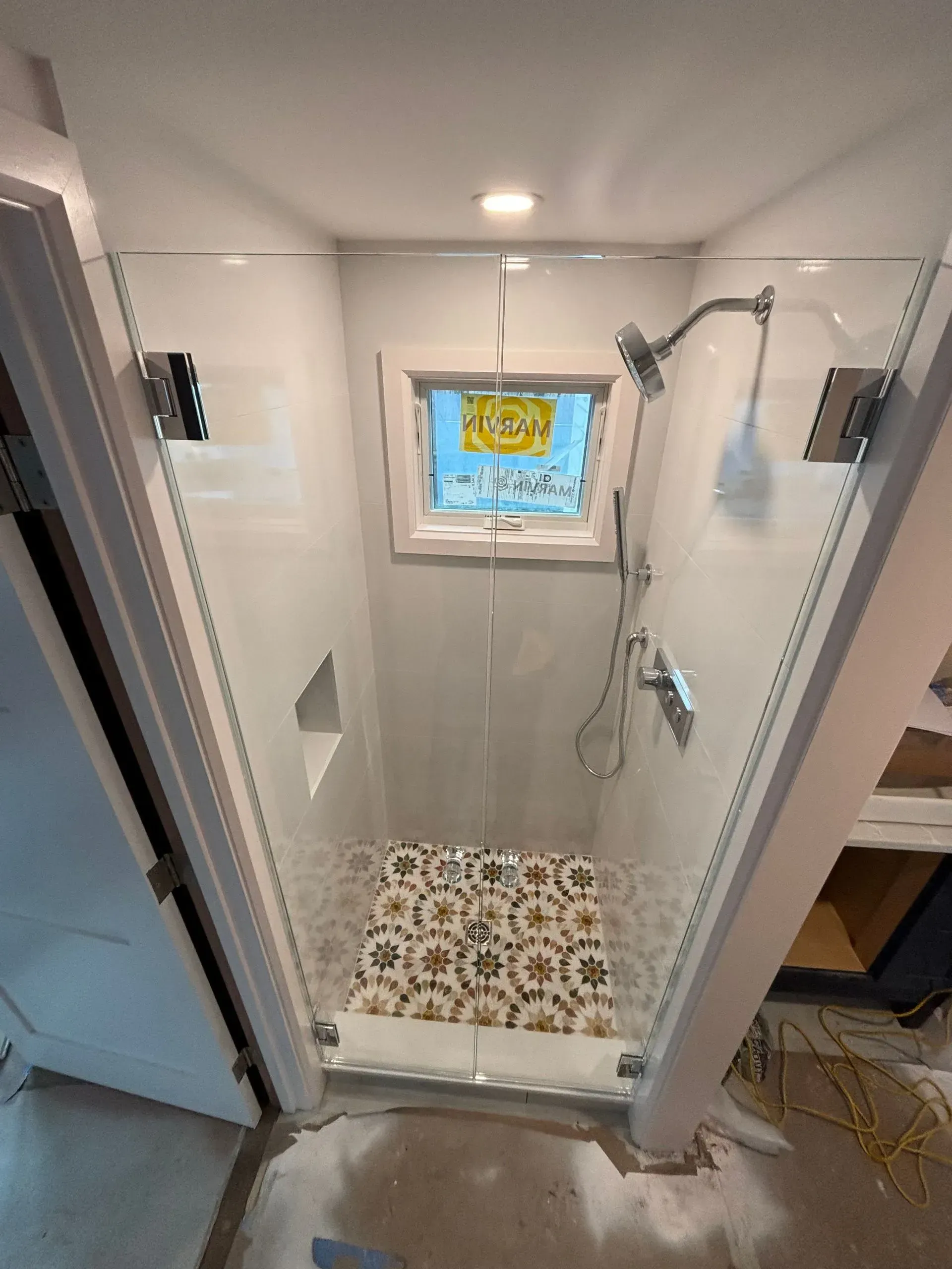 A newly installed shower with glass doors, white walls, and patterned tile floor; a window at the back.