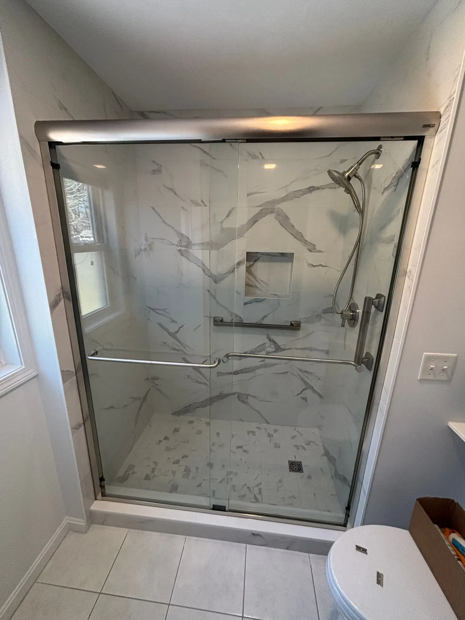 Modern shower with marble-look walls, glass doors, and chrome hardware. Includes a grab bar, built-in shelf, and overhead showerhead.
