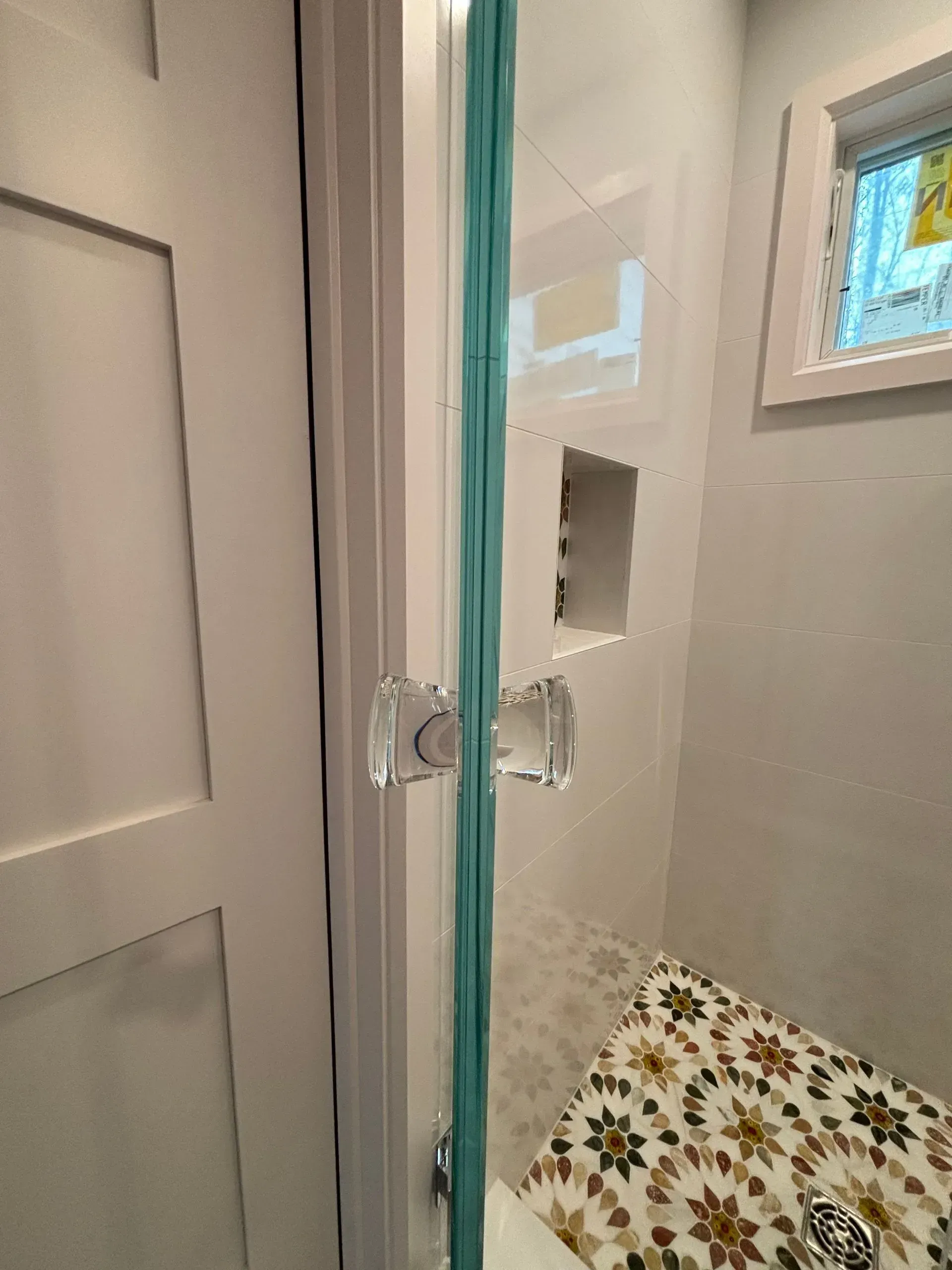 Shower door with clear glass, a glass handle, and a white-tiled wall with a mosaic bench.