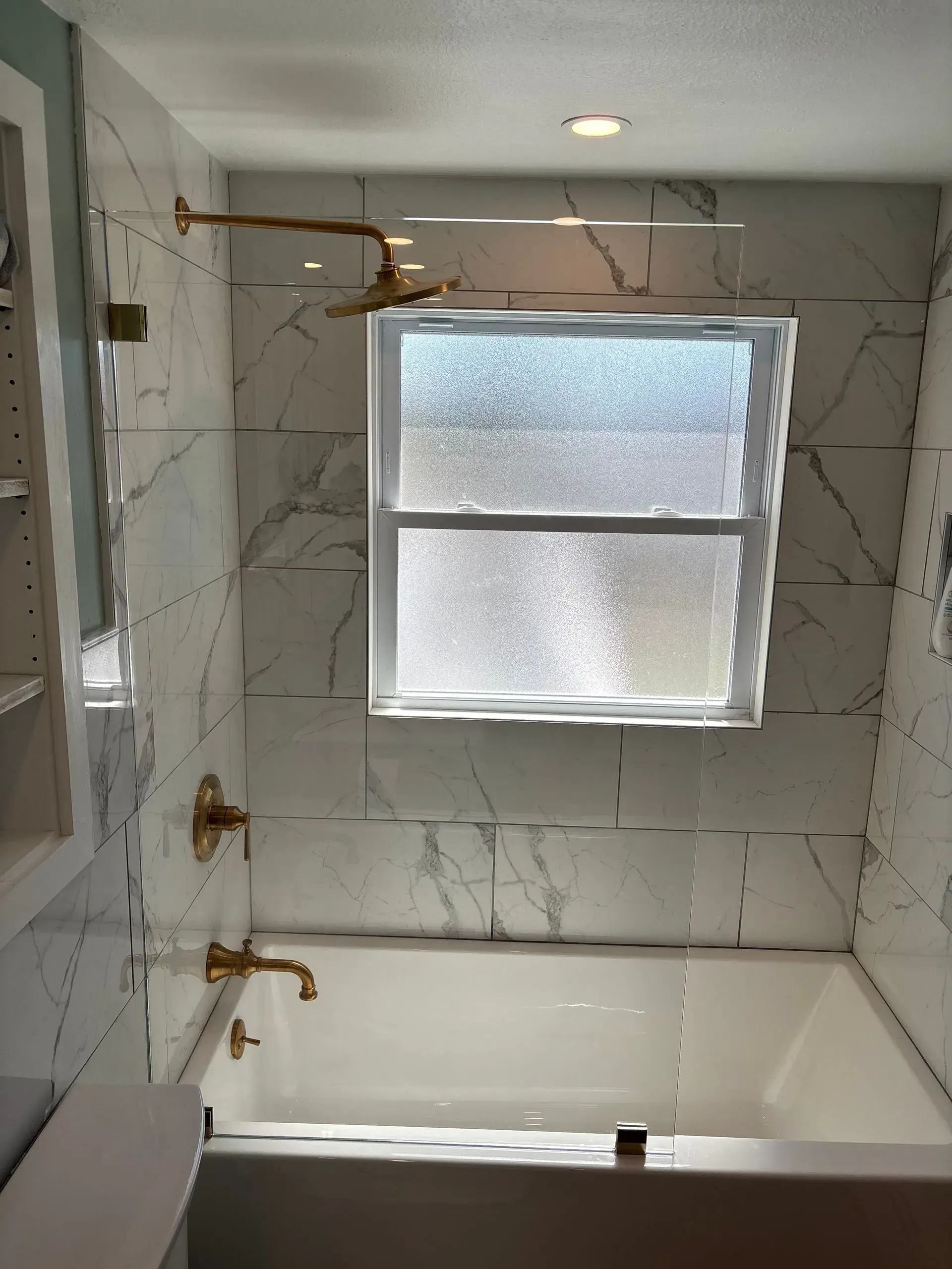 Bathroom with white marble tile, gold fixtures, tub under a frosted window.
