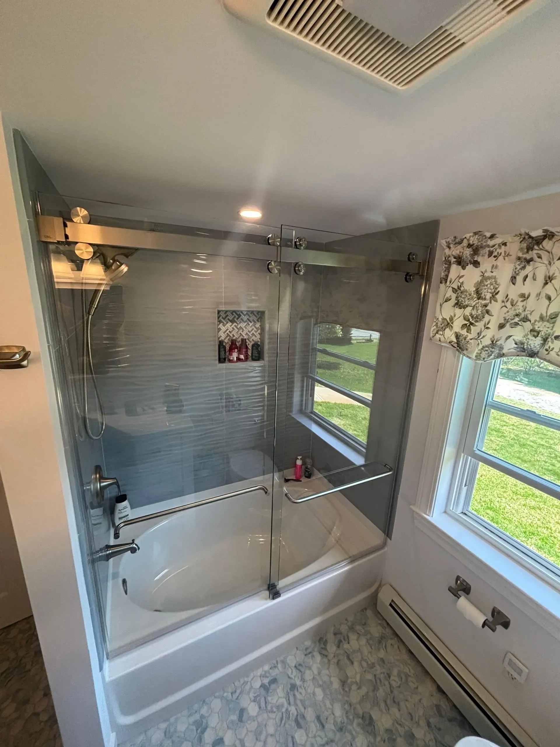 Bathroom with glass shower enclosure over a white tub, window with floral valance, gray tile floor.