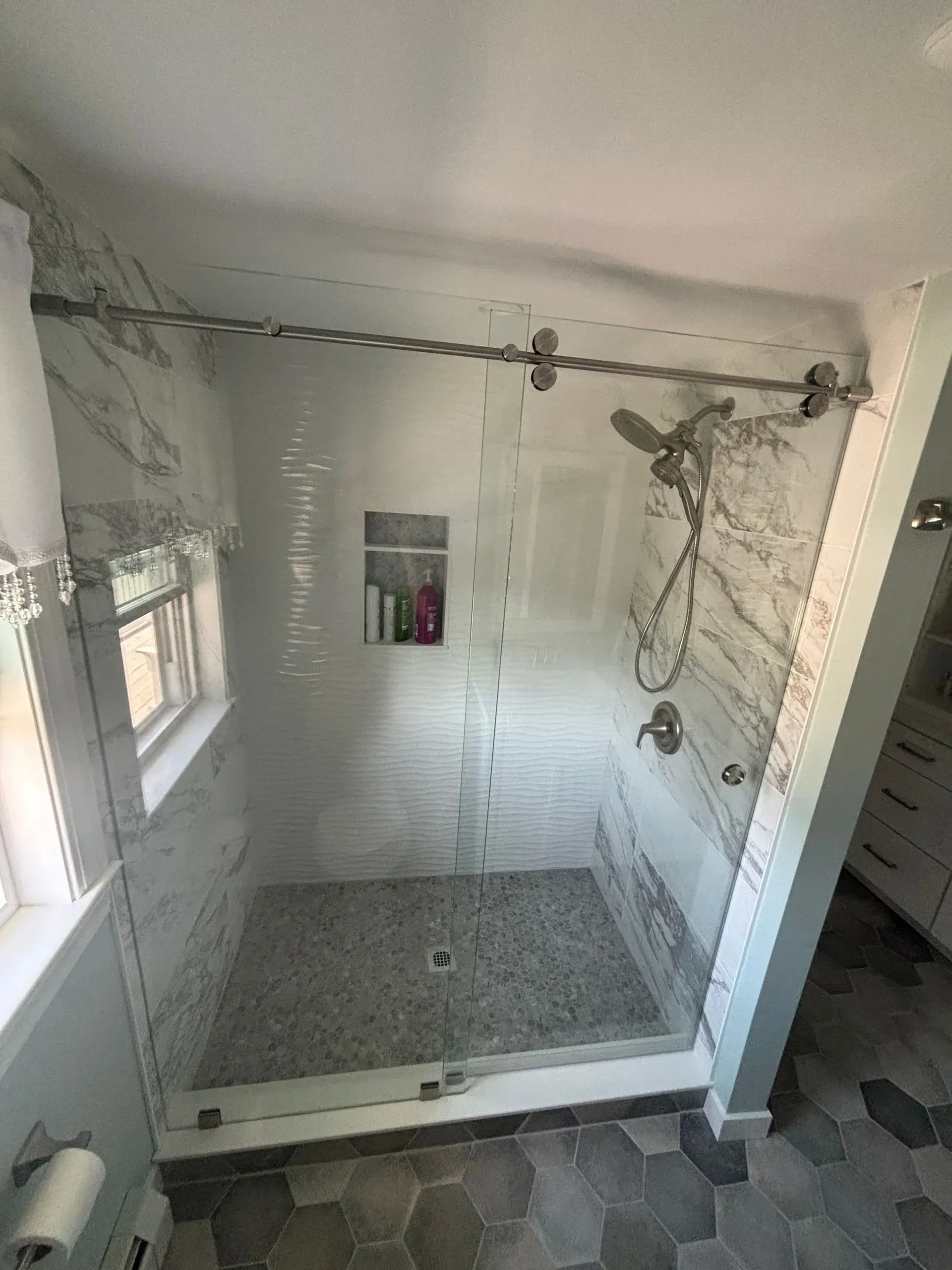 Glass shower with sliding door and marble-patterned walls, pebble shower floor, and overhead shower head.