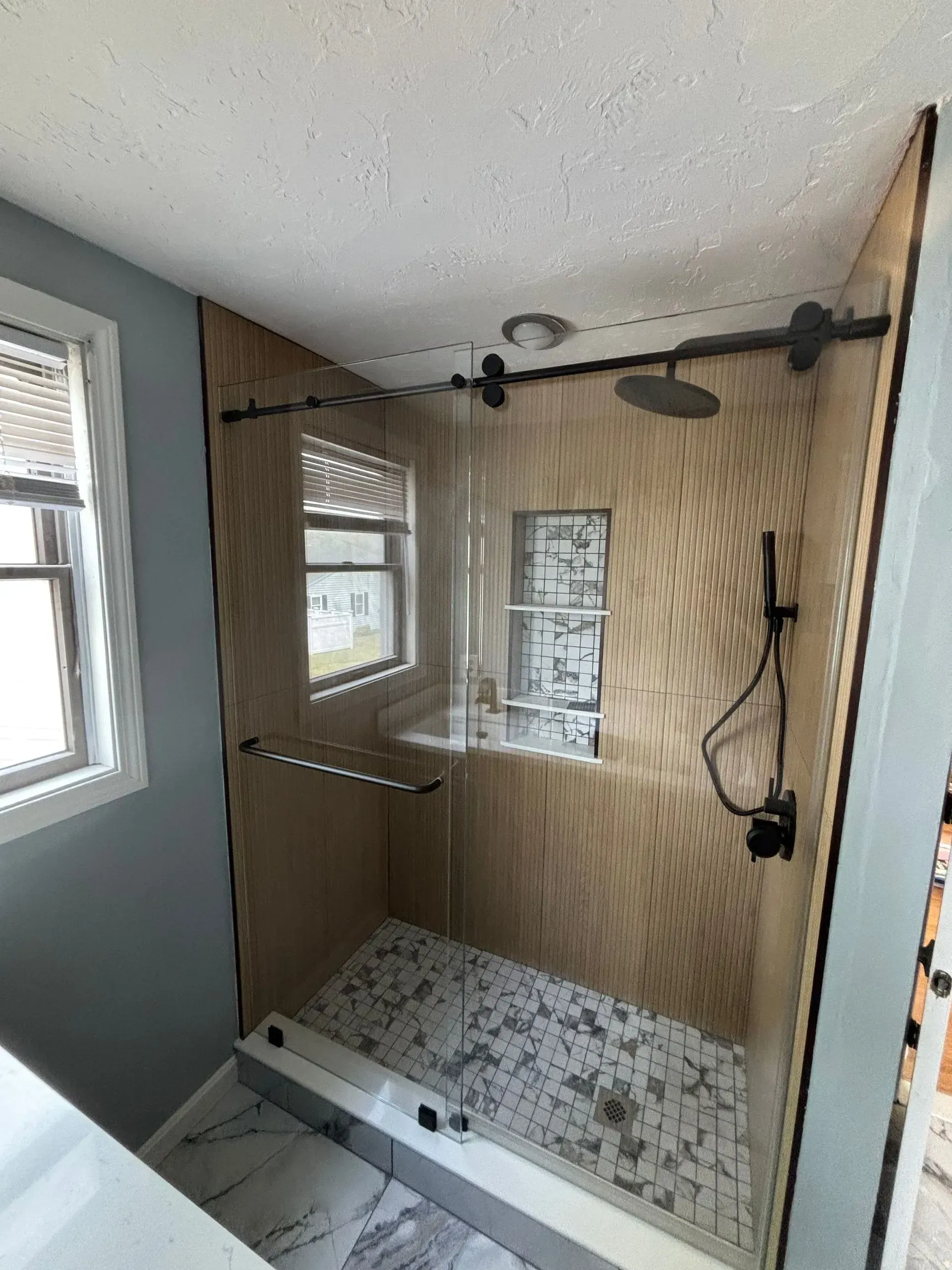 Shower with glass doors, light wood paneling, black fixtures, and patterned floor tiles next to a window.