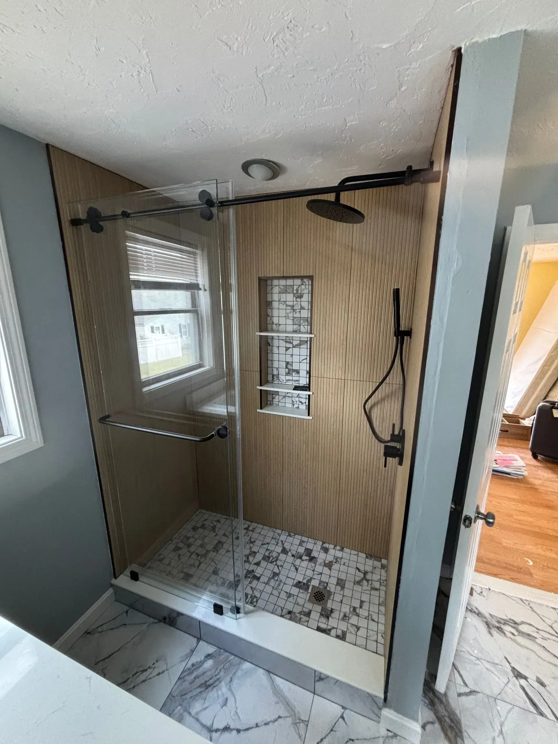 Modern shower with glass door, black fixtures, light wood paneling, and mosaic tile flooring.