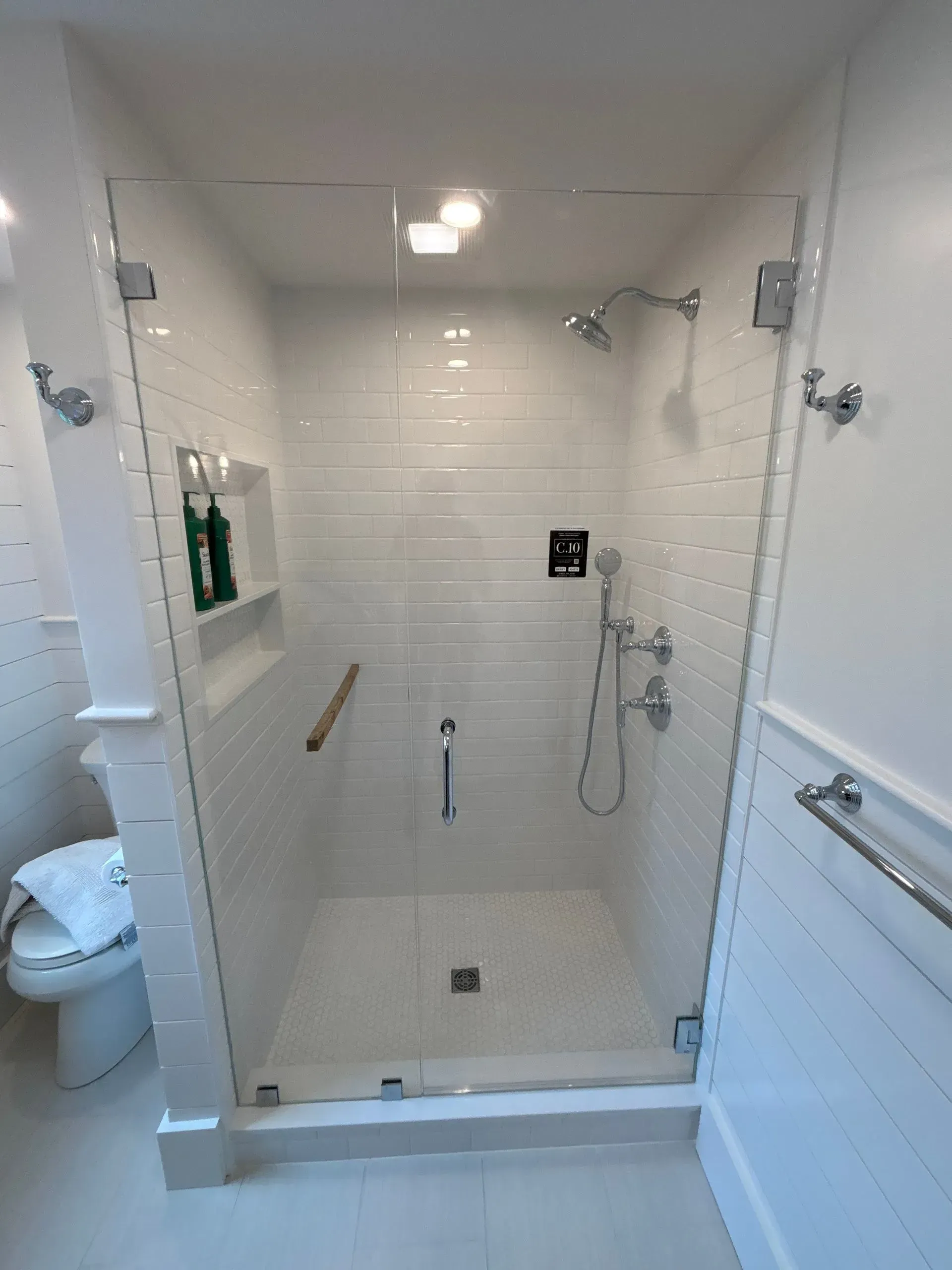 White tiled shower with glass doors, grab bar, and showerheads, next to a toilet.