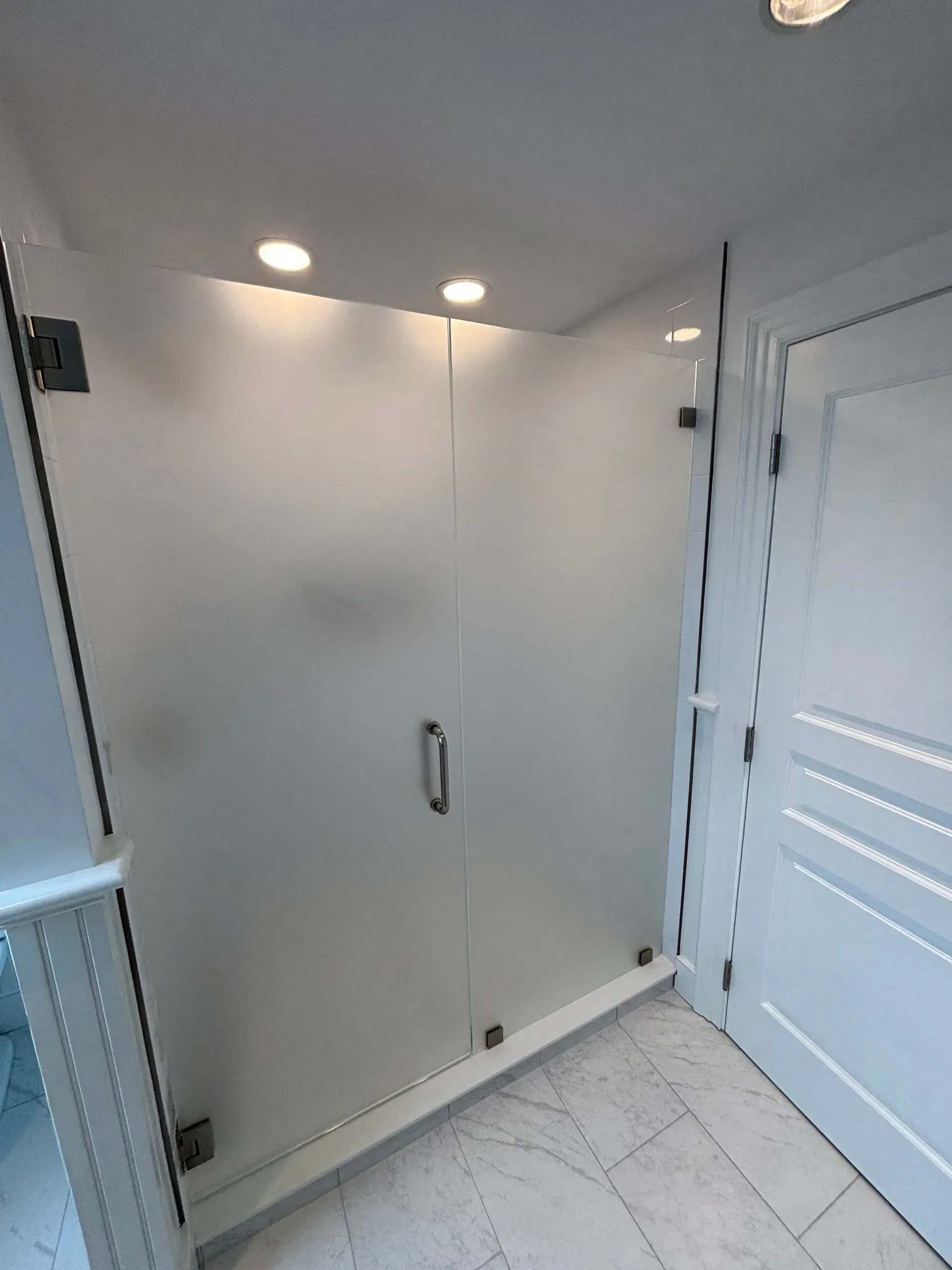 Frosted glass shower enclosure with a door and handle in a white bathroom, lit by overhead lights.