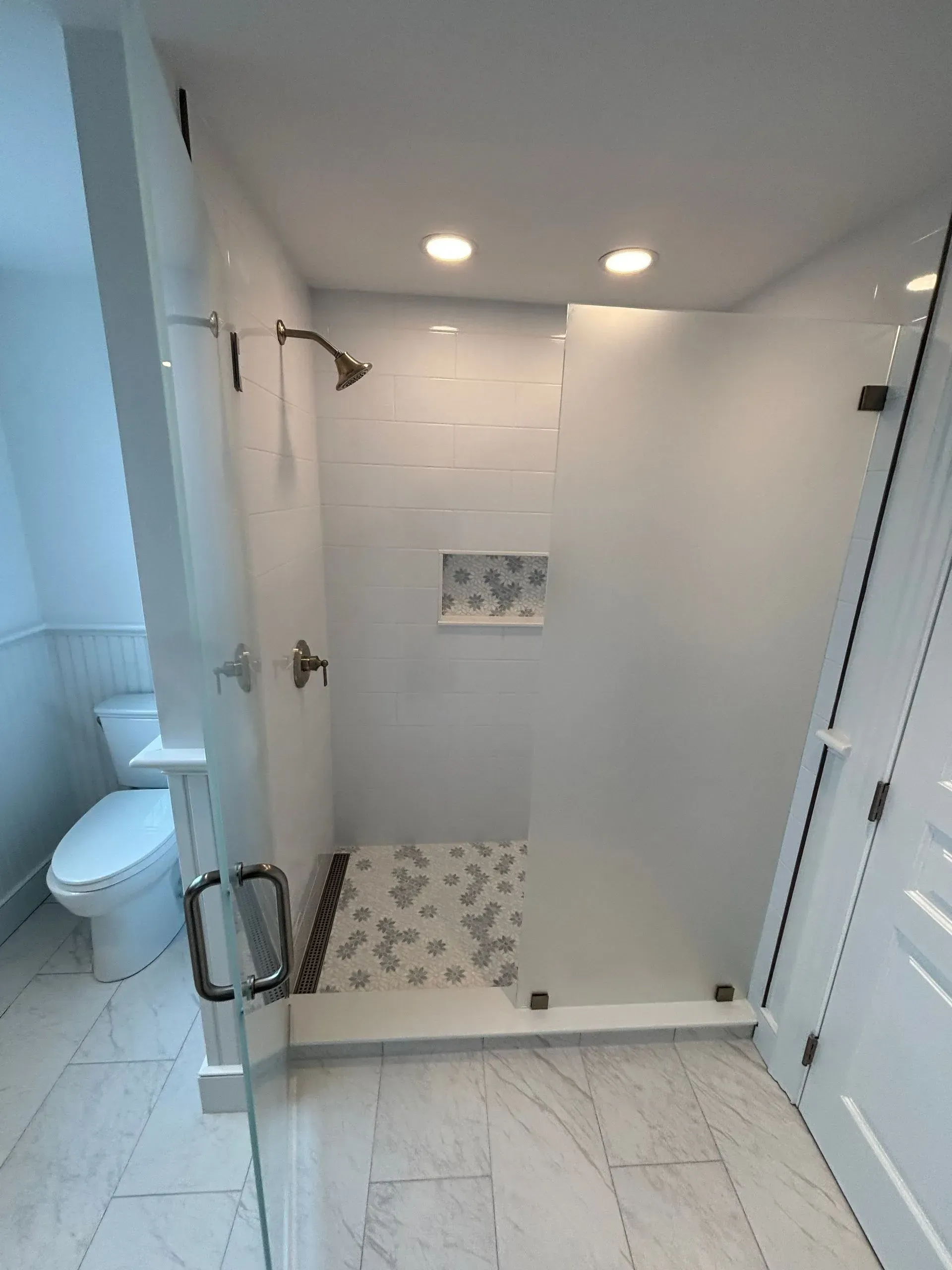 Modern bathroom with glass shower, white tile, gray floor, toilet, and white door.