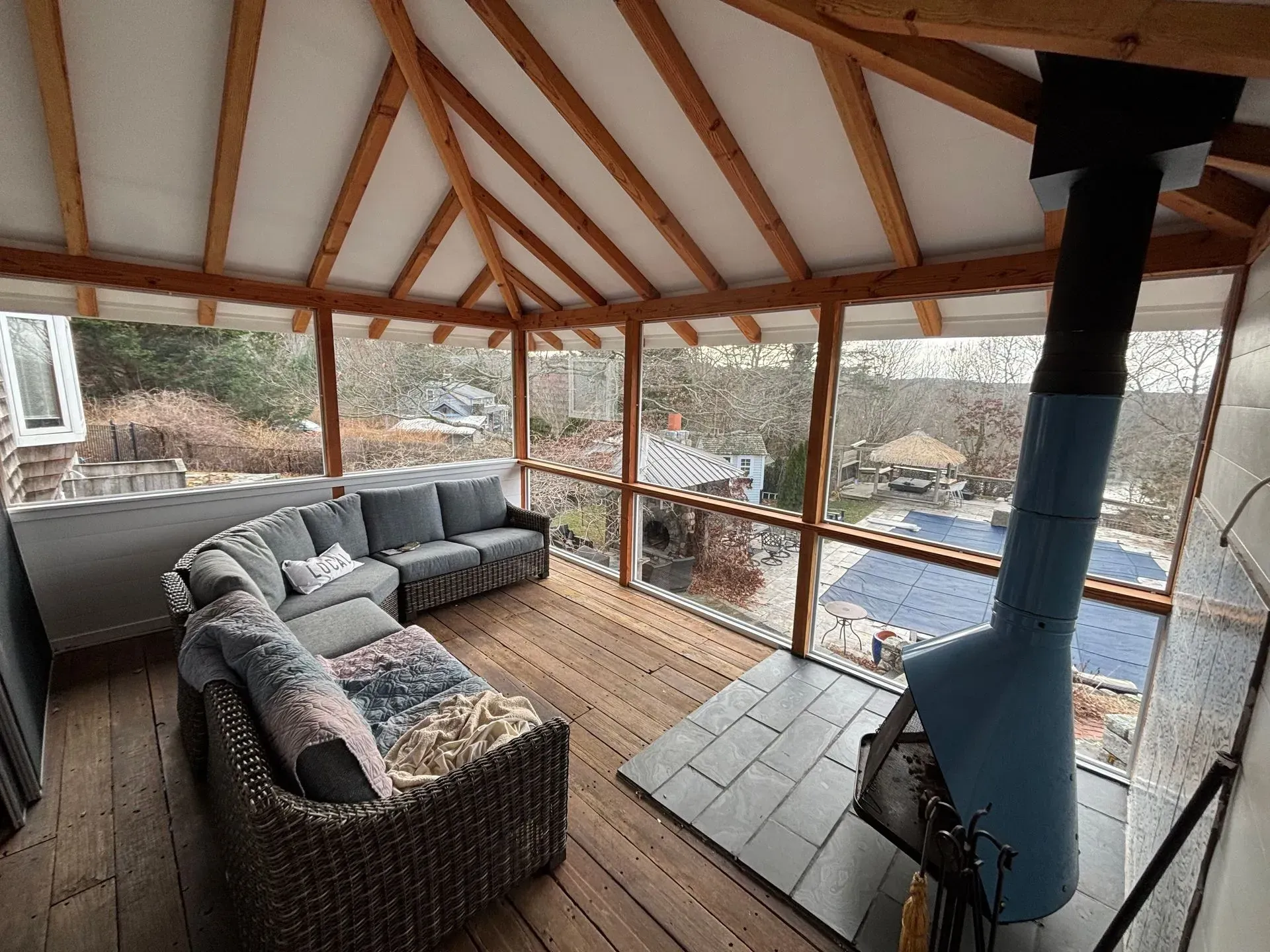 Cozy outdoor seating area with a fireplace. Gray sectional sofa and blue wood stove in a screened-in porch.