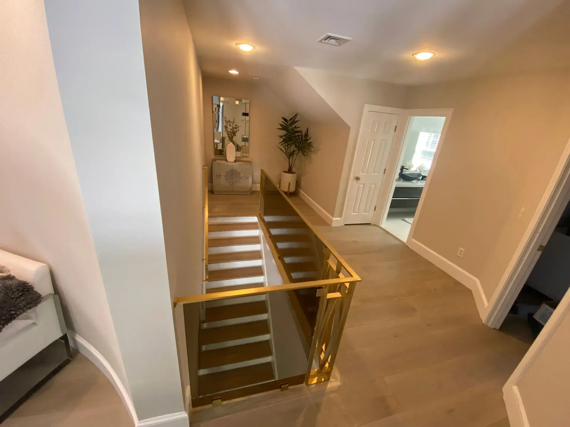 Hallway with gold railing, stairs, neutral walls, and hardwood floors. A mirror and potted plant decorate.