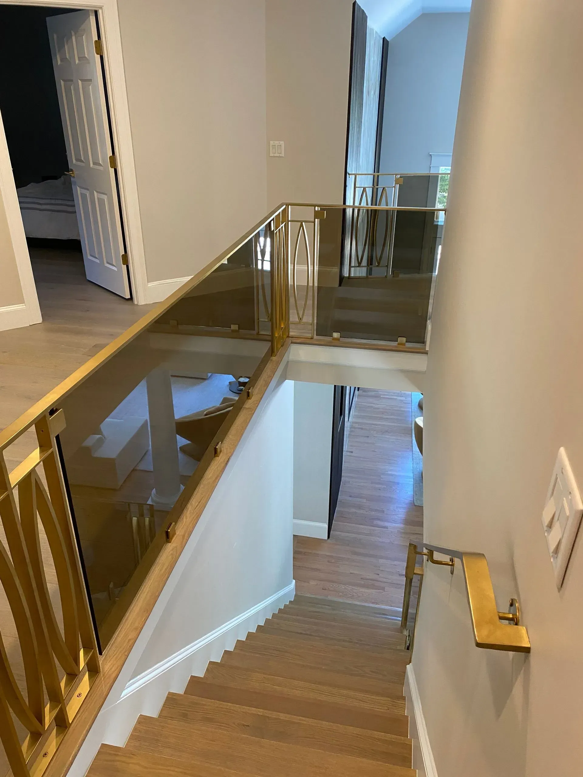 A staircase with a gold railing and glass panels leading down, with a view of a hallway.