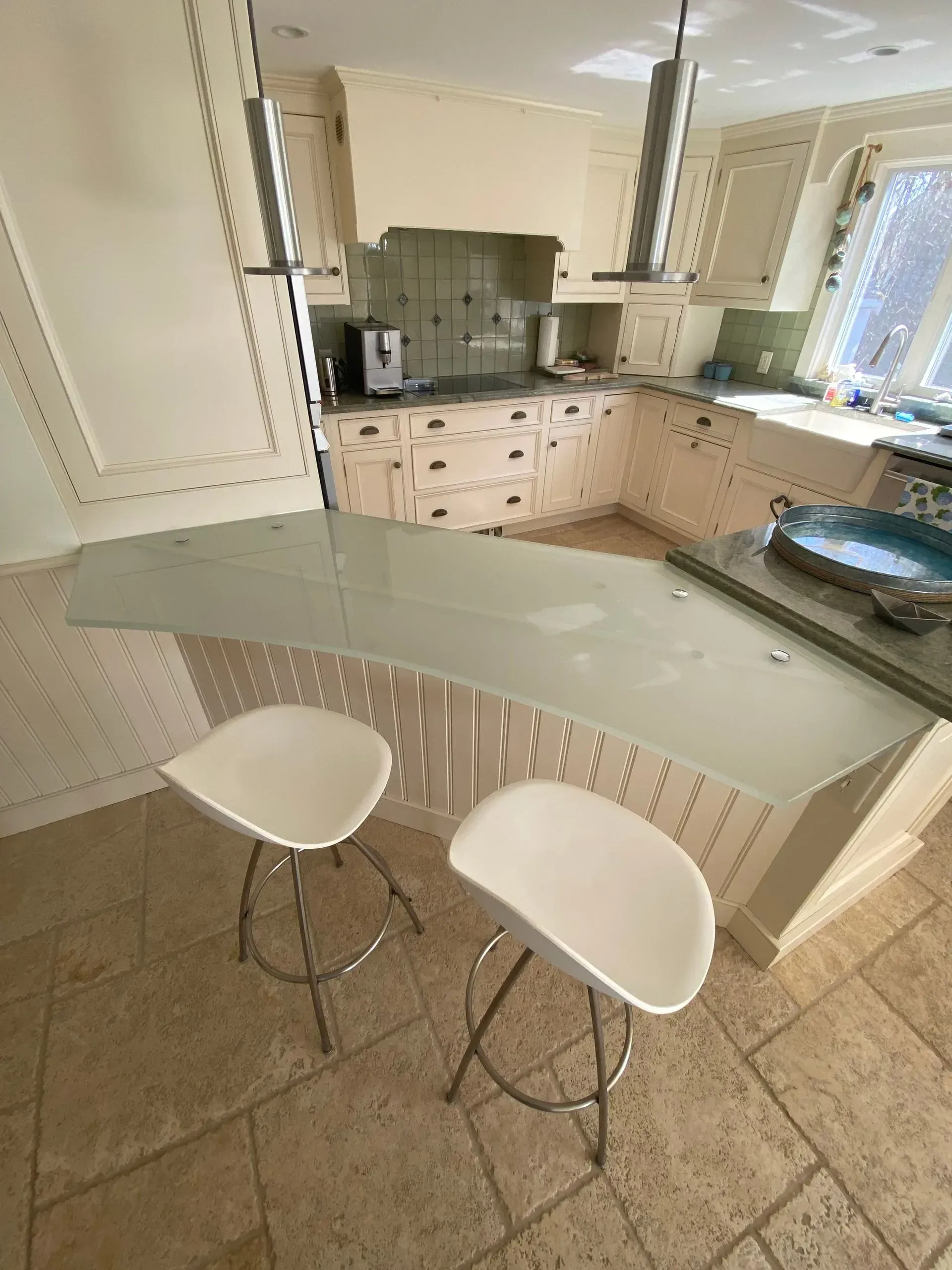 Two white bar stools at a light-colored kitchen island. Cream cabinets, tiled floor, and glass backsplash.