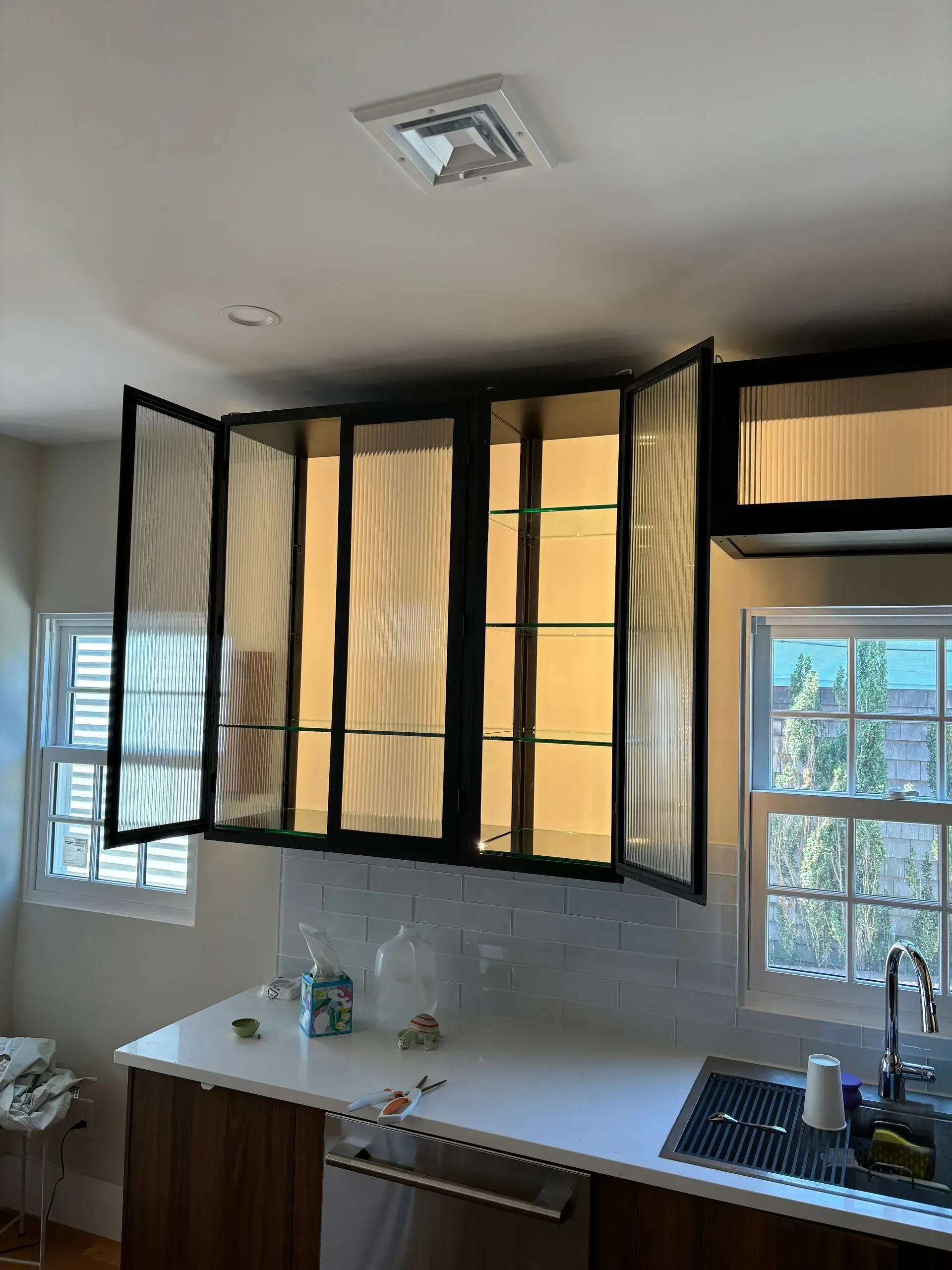 Kitchen with black-framed cabinets with glass doors over a white countertop and sink; two windows with light entering.