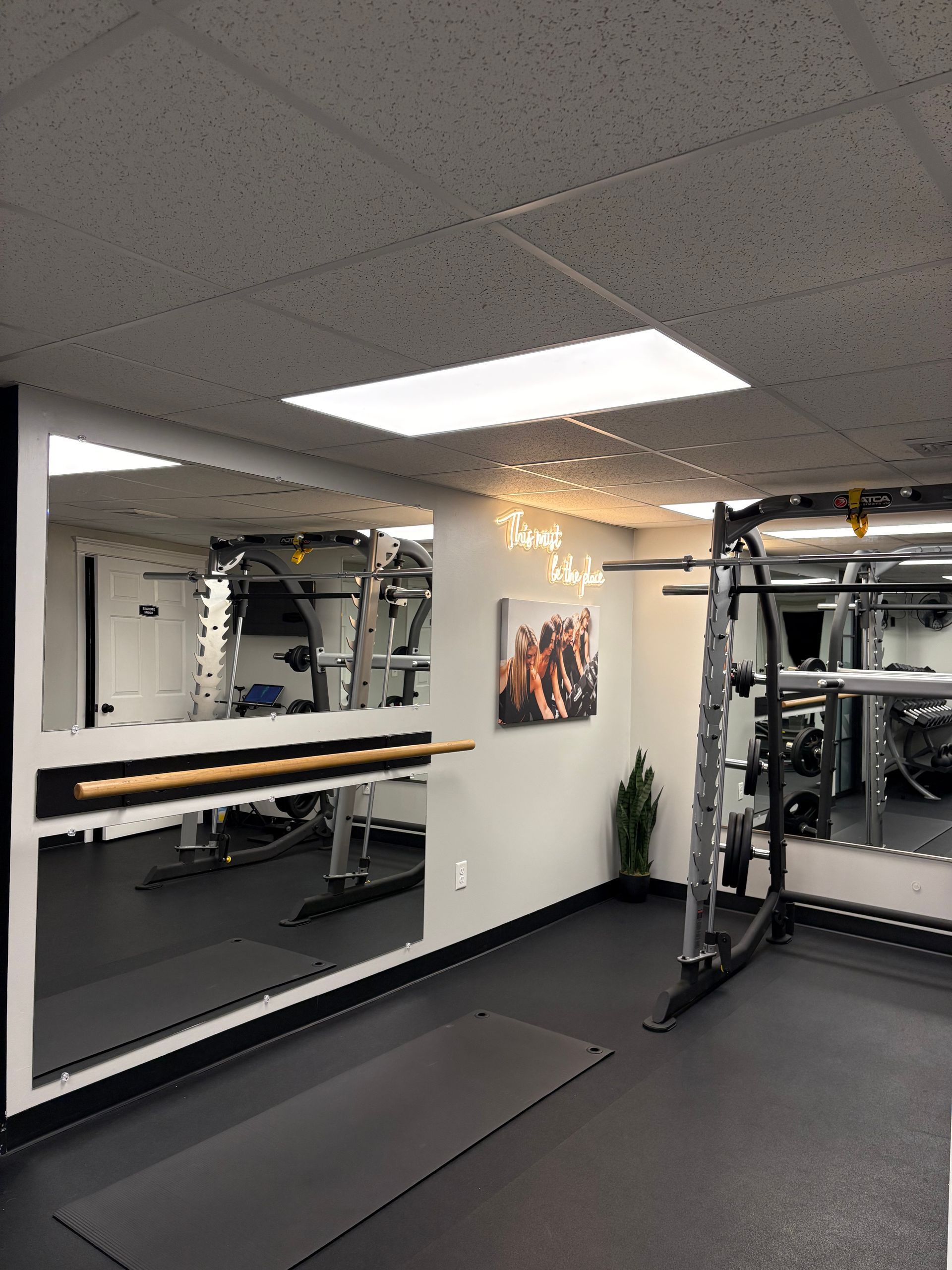 Gym with mirror wall, weights, and equipment. Dark floor, light ceiling, inspirational art.