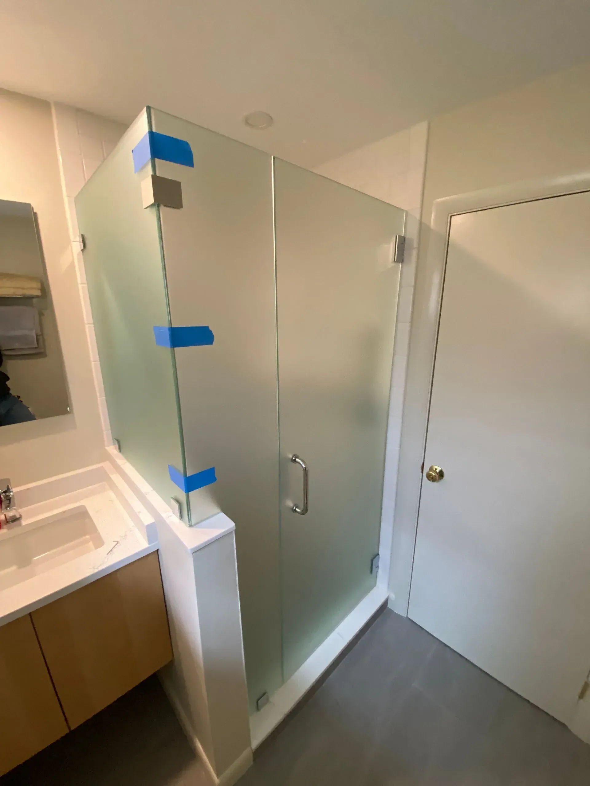 Frosted glass shower enclosure in a bathroom; white walls, gray tile floor, sink and door visible.