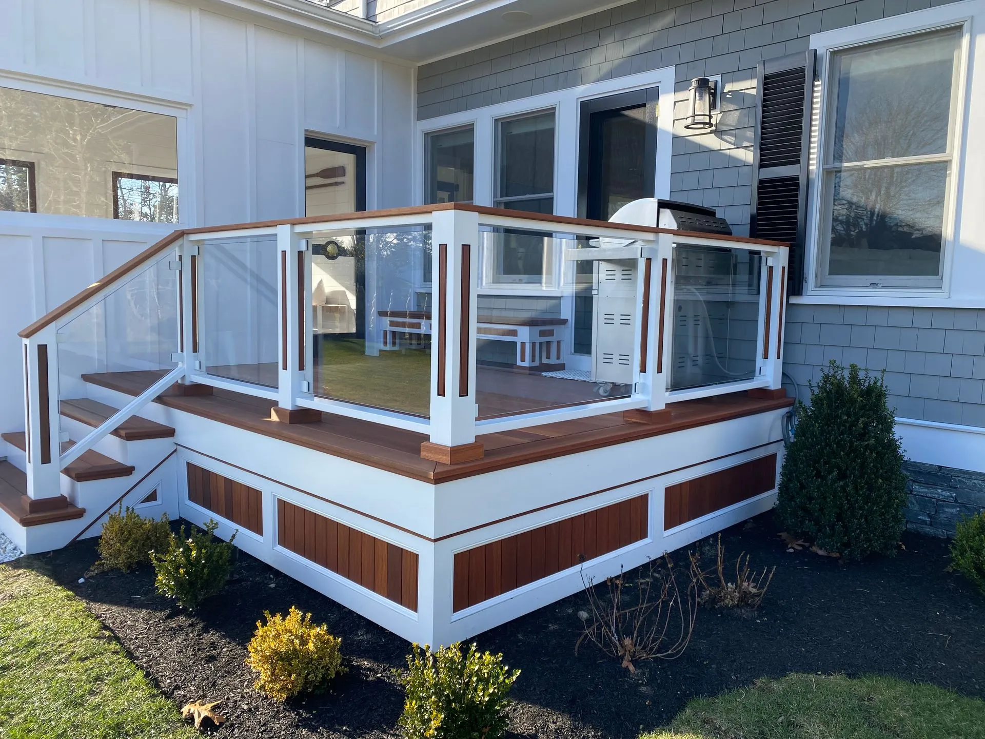 Deck with glass railings, brown and white trim, stairs leading up, shrubs and grass surrounding.