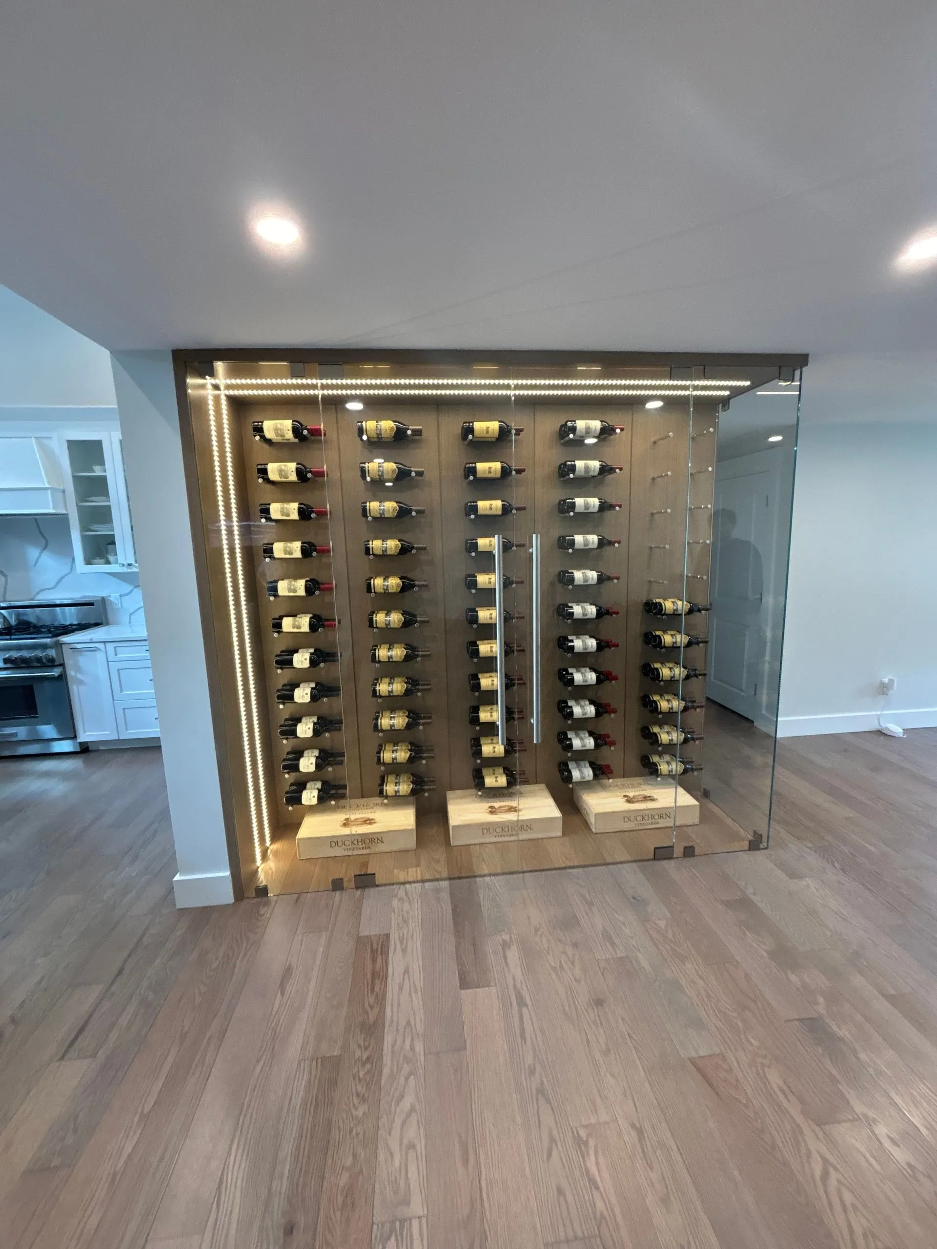 Wine rack built into a wall with bottles, wooden flooring, and kitchen visible in the background.