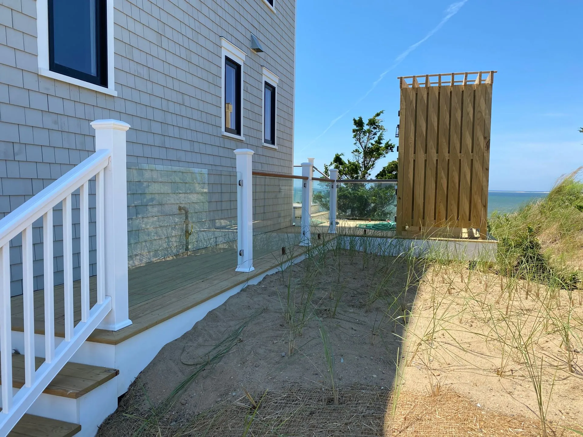 Beach house with glass fence, wooden shower enclosure, and stairs leading down. Sunny day.