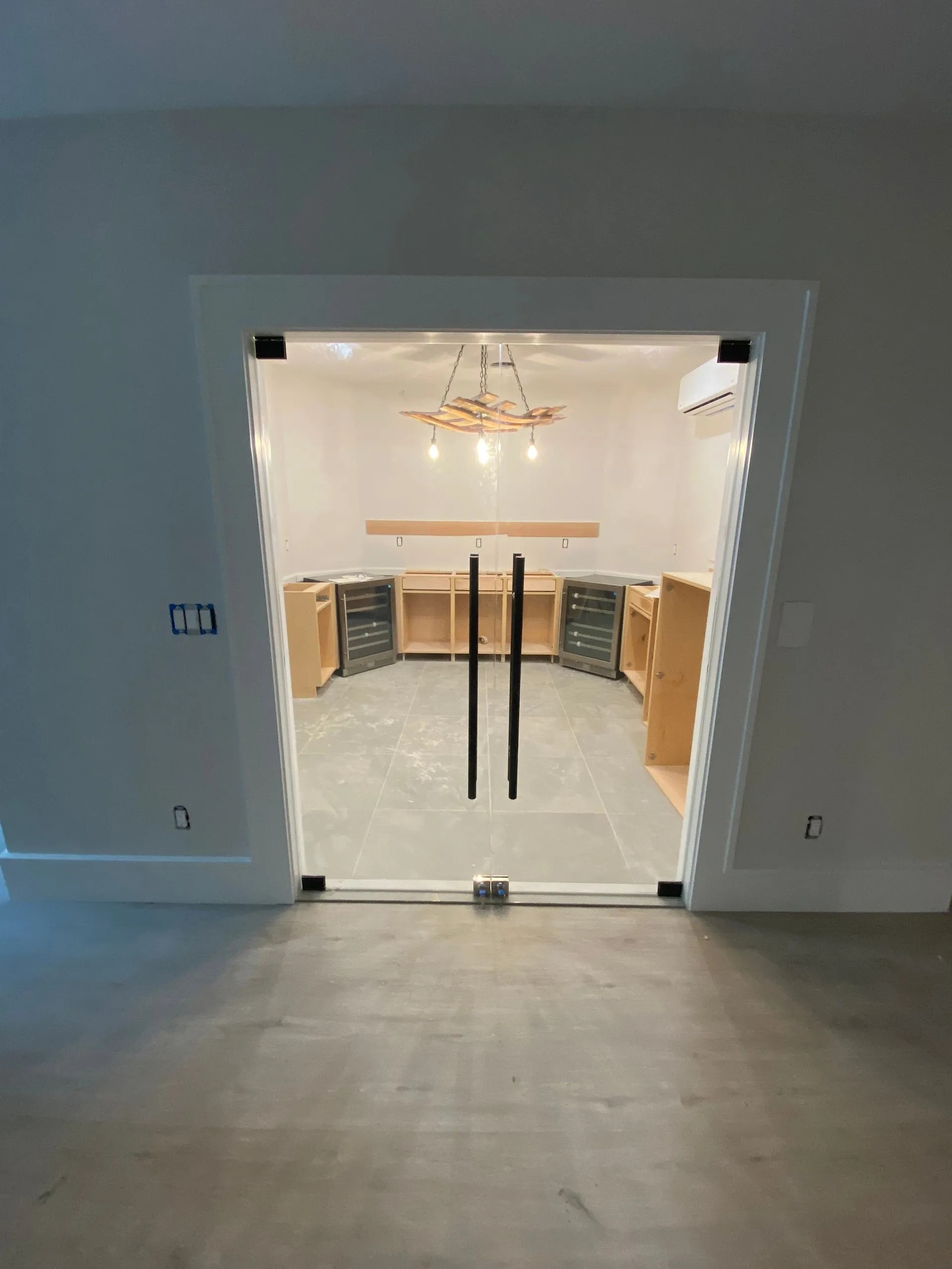 Glass door entrance to a wine room with unfinished cabinets, light fixtures and AC unit.