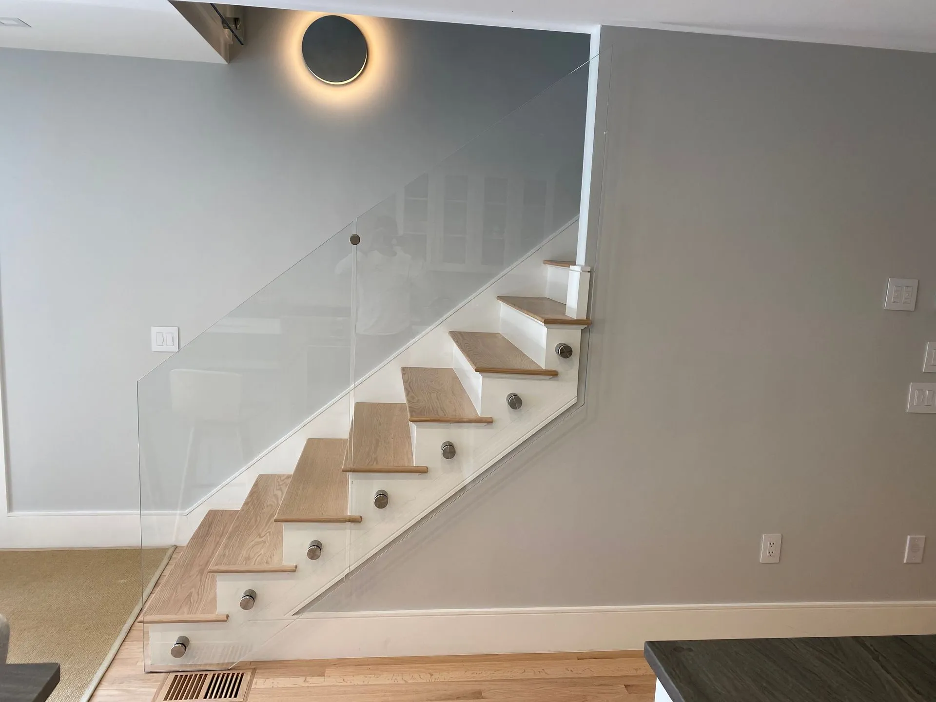 Staircase with glass railing, light wood steps, grey walls, and a round wall light.
