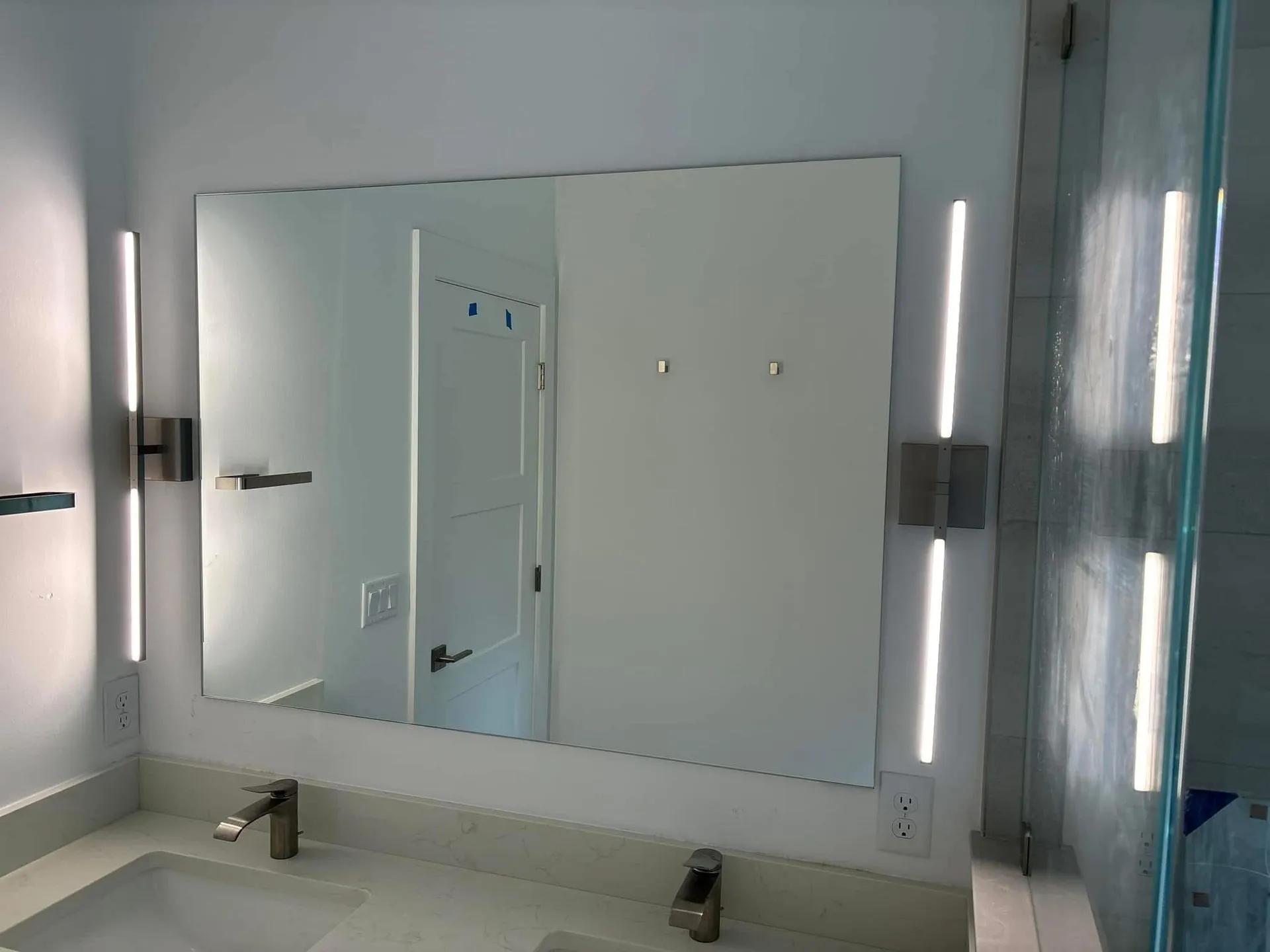 Bathroom mirror with vertical LED lights, two sinks, and a glass shower door.