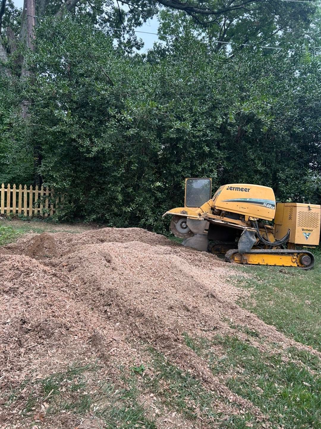 Yellow wood chipper beside a large pile of mulch in a wooded yard with a fence and trees.