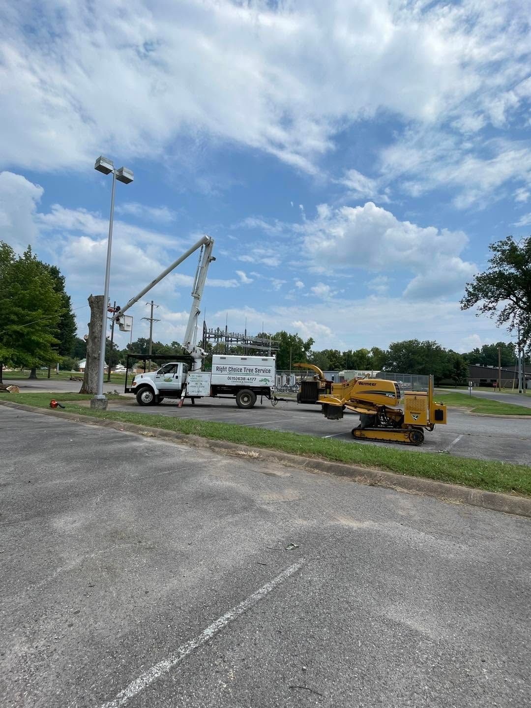 Utility trucks and a yellow lift parked beside a road under a cloudy sky, with trees and a grassy lot nearby.