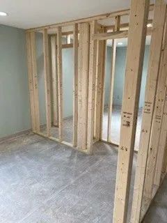 Framed interior wall under construction in an unfinished room with gray flooring and exposed wood studs.