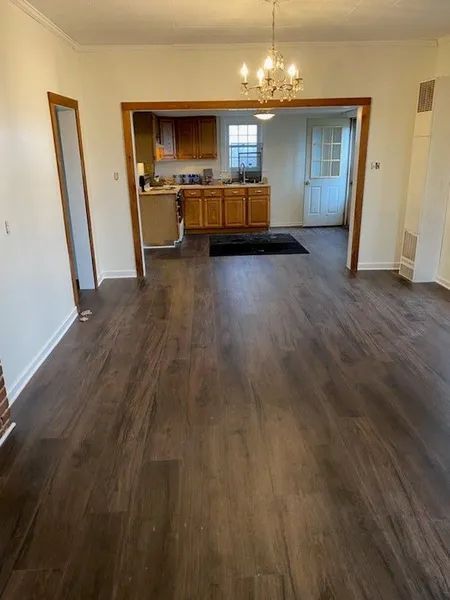 Empty living room with wood-look floors, chandelier, and a view into a kitchen with wood cabinets.
