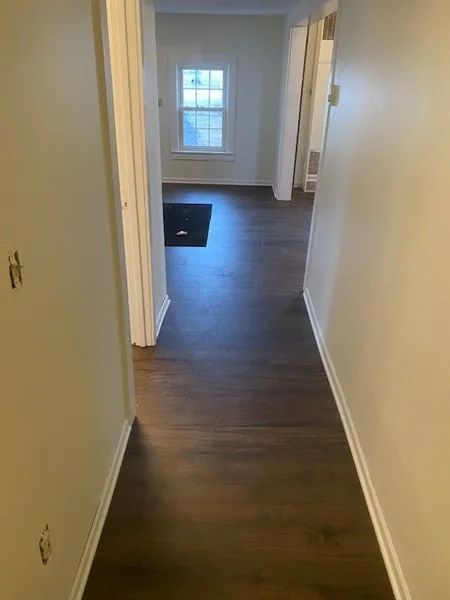 Empty hallway with dark wood floors, white walls, and a window at the far end.