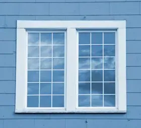 White-framed double window on a blue clapboard wall, reflecting cloudy sky.