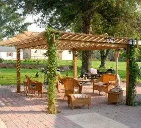 Wooden pergola over a brick patio with wicker chairs, tables, and vines in a grassy park setting