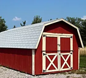 Red barn with white trim and double doors under a gray shingled roof on a gravel lot